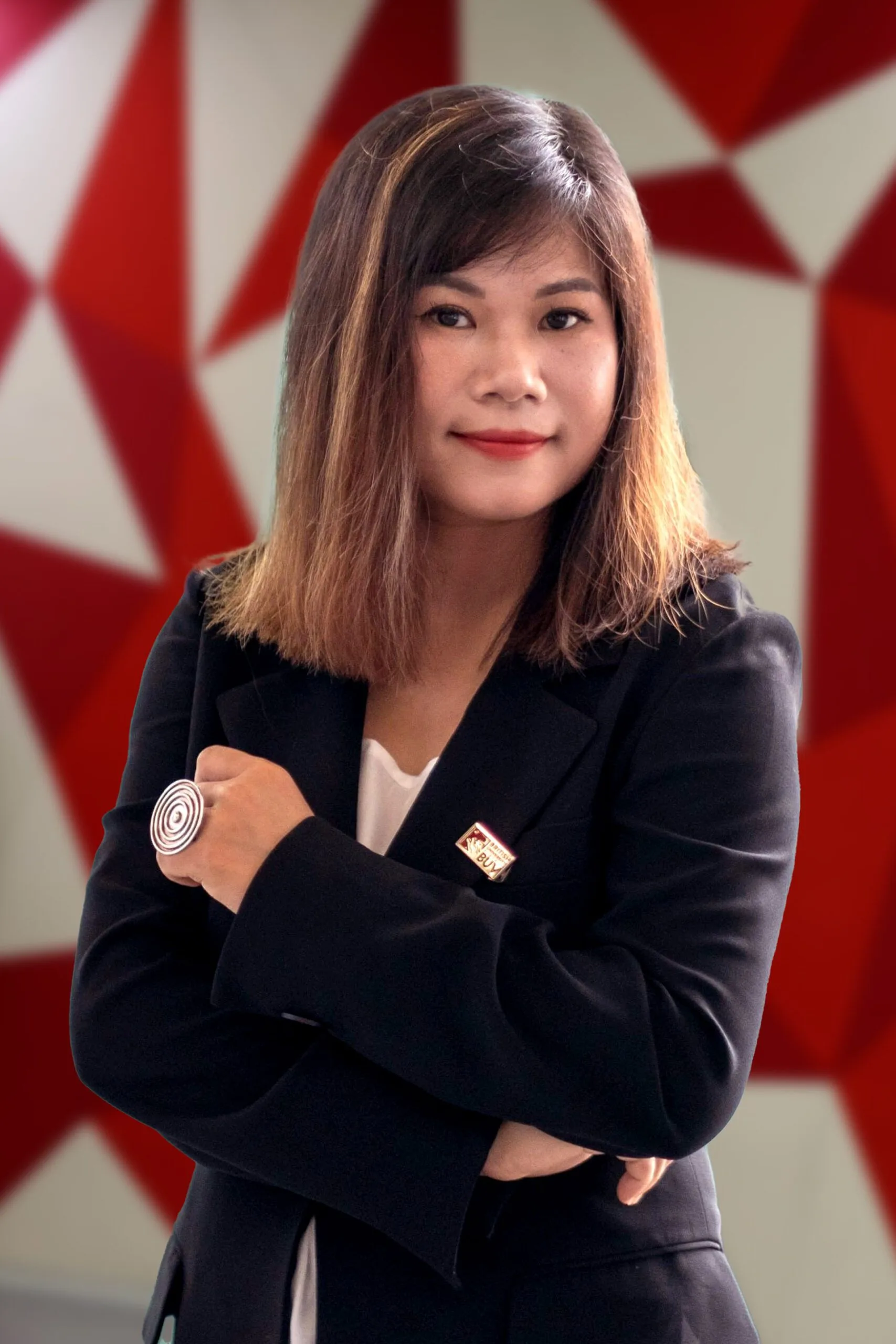 A portrait of a woman in a black blazer with arms crossed, standing in front of a red geometric backdrop.