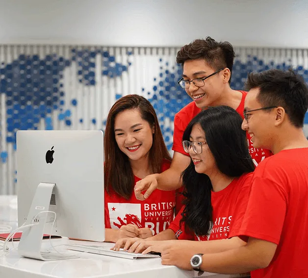 Four BUV students in red university T-shirts smile and collaborate around an Apple desktop computer.