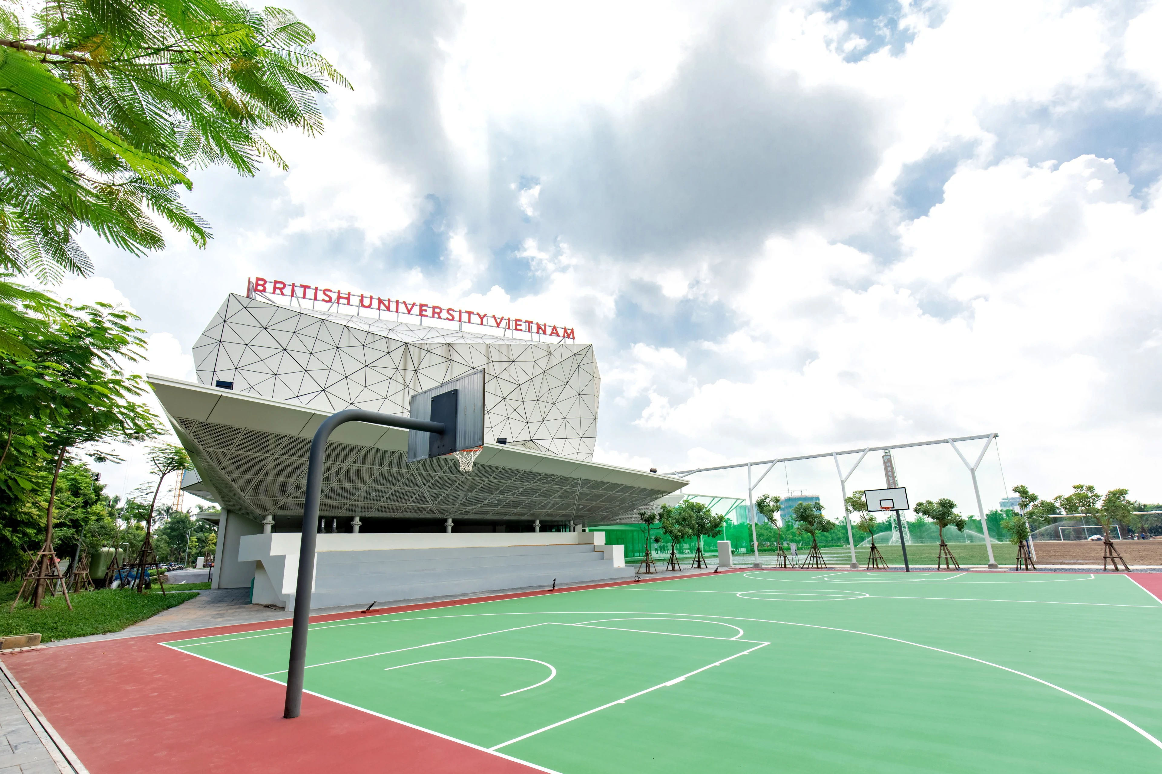 British University Vietnam campus with an outdoor basketball court, modern geometric building, greenery, and a partly cloudy sky.