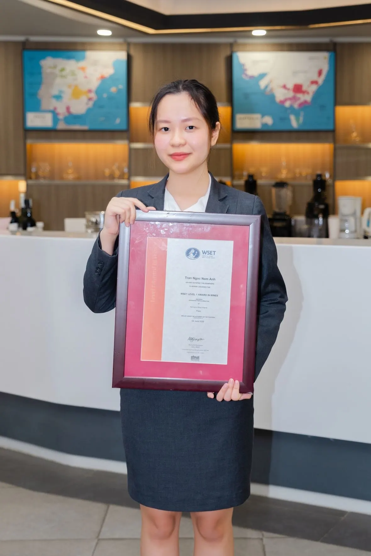 A woman in a dark blazer and skirt stands at a reception desk, holding a framed certificate with a red border; colorful world maps are displayed on the wall behind her.