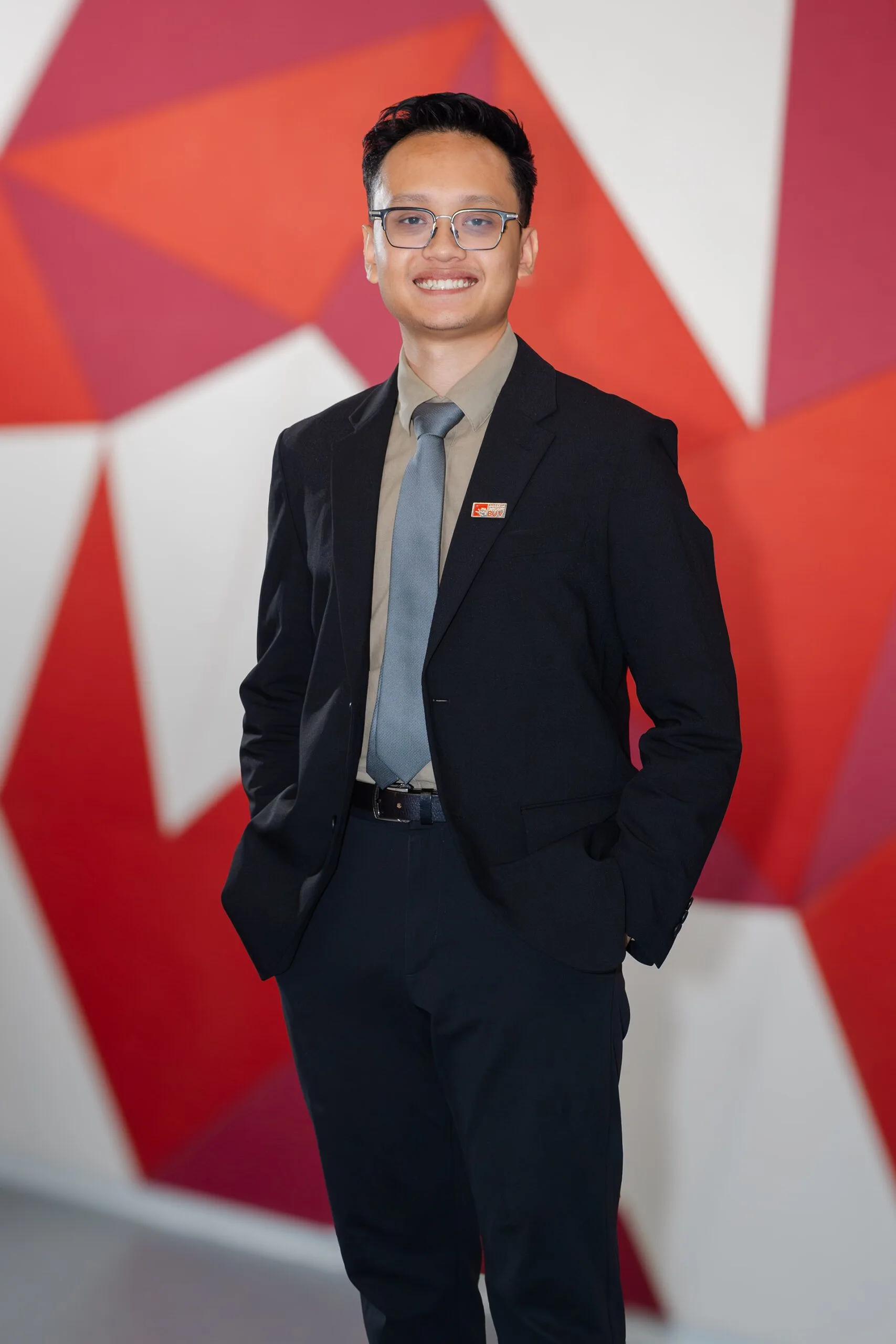 A man in a dark suit and tie stands smiling for a formal portrait against a red-and-white geometric background.