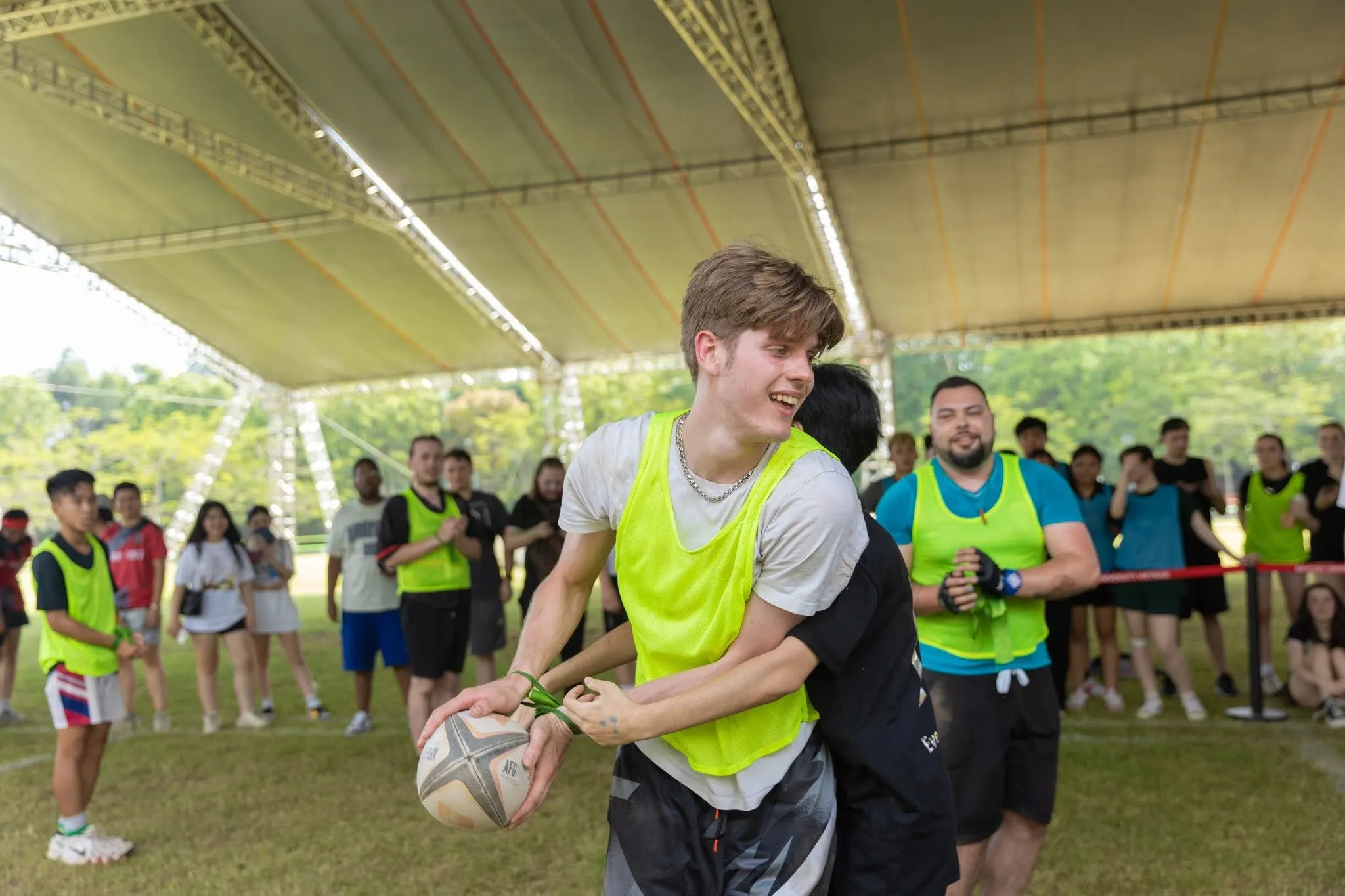 BUV students play rugby under a covered sports field, wearing fluorescent training bibs.