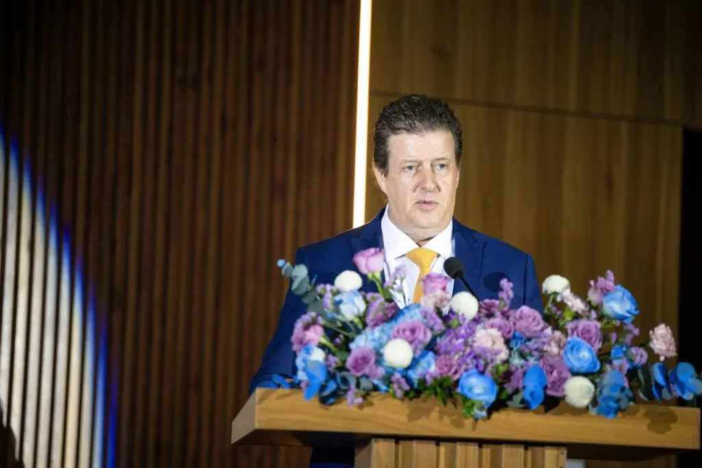 A man in a blue suit speaks at a wooden podium adorned with a large arrangement of colorful flowers.