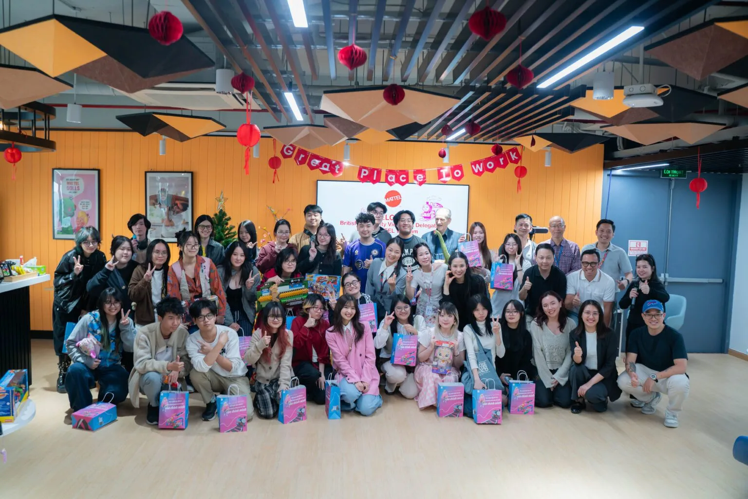 Group photo of students and staff celebrating beneath red lanterns and “Good Place to Work” decorations, with colorful gift bags arranged in front.