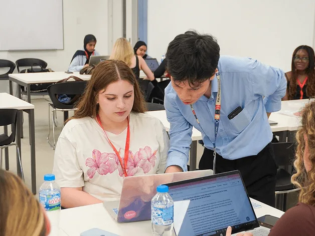 Two students collaborate in a classroom, reviewing information on a laptop and tablet while other students work in the background.