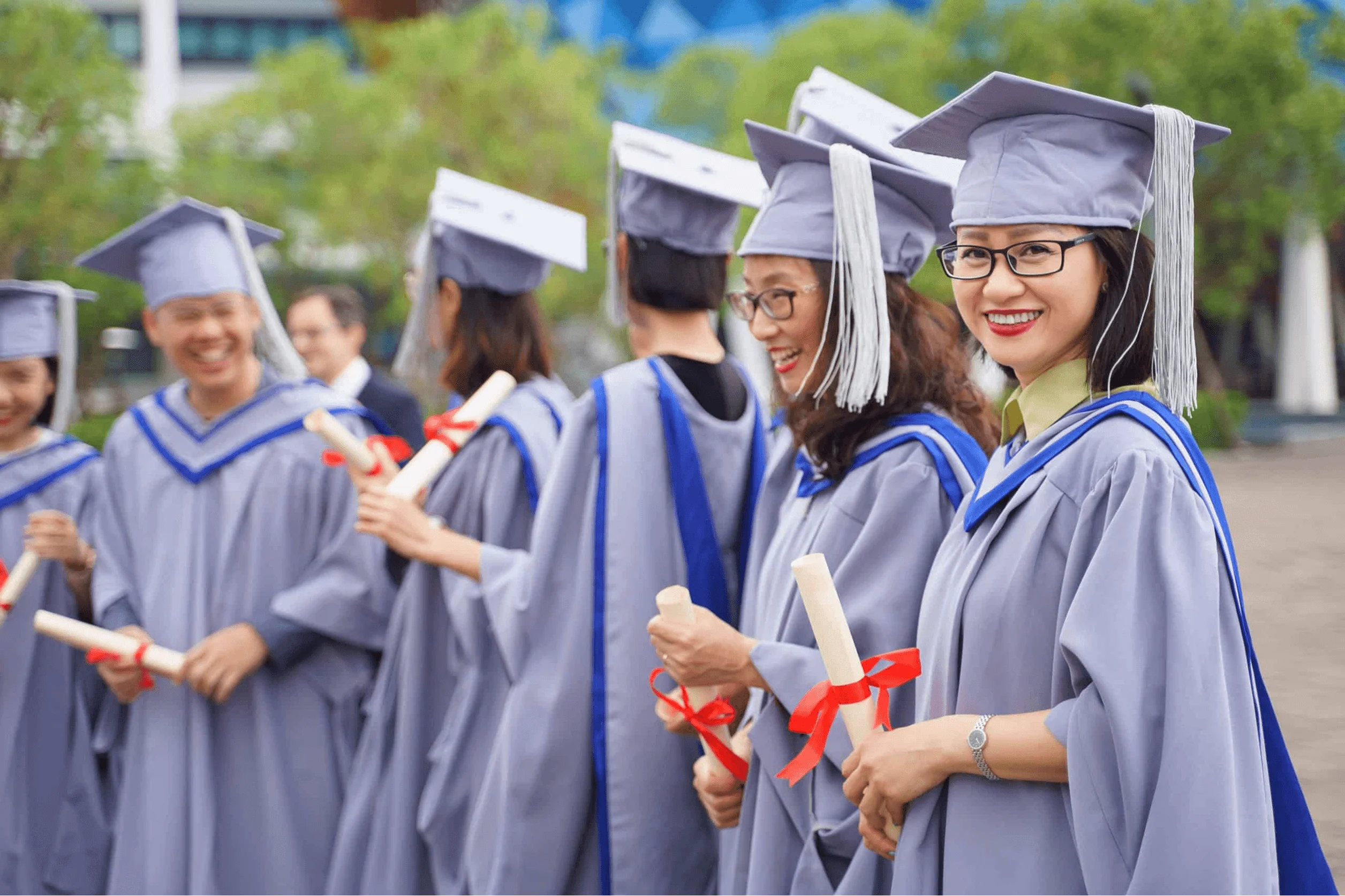 Smiling university graduates standing in a row, wearing grey caps and gowns while holding tied diplomas.