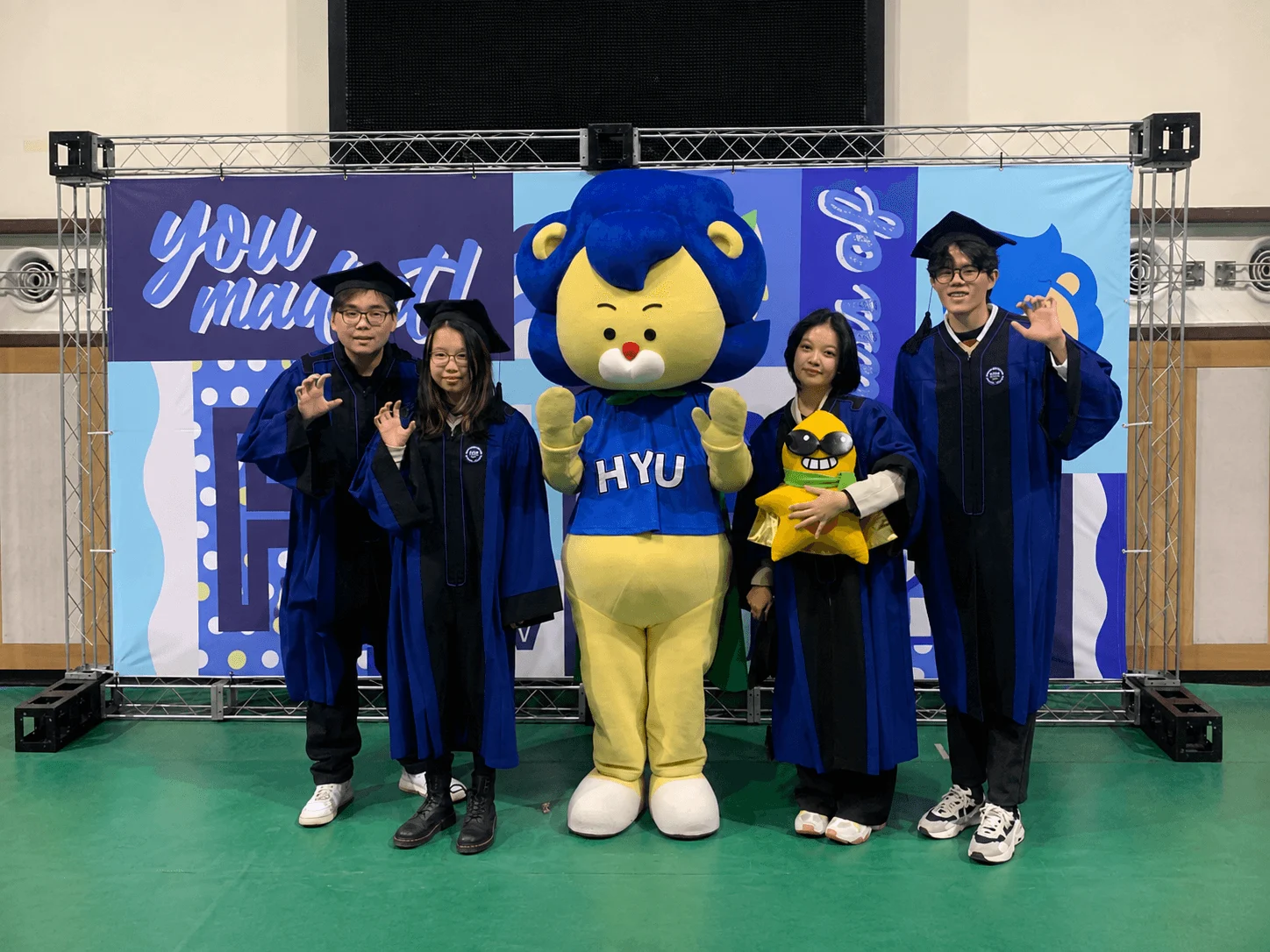 Four students in blue academic gowns pose with the HYU lion mascot at an exchange event.