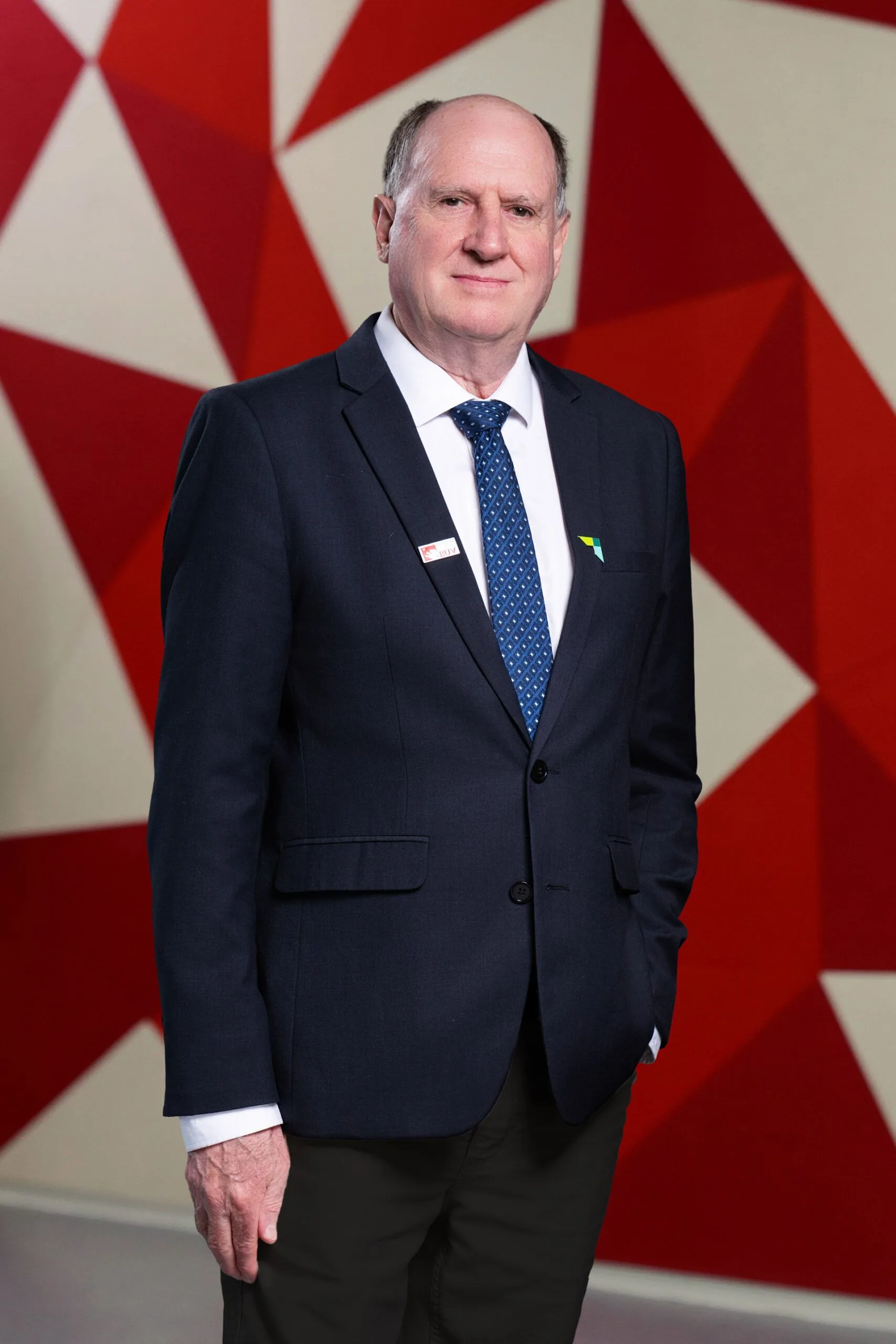 Portrait of Mark Wheaton standing and smiling in a dark suit with a blue tie in front of a red and white Union Jack backdrop.