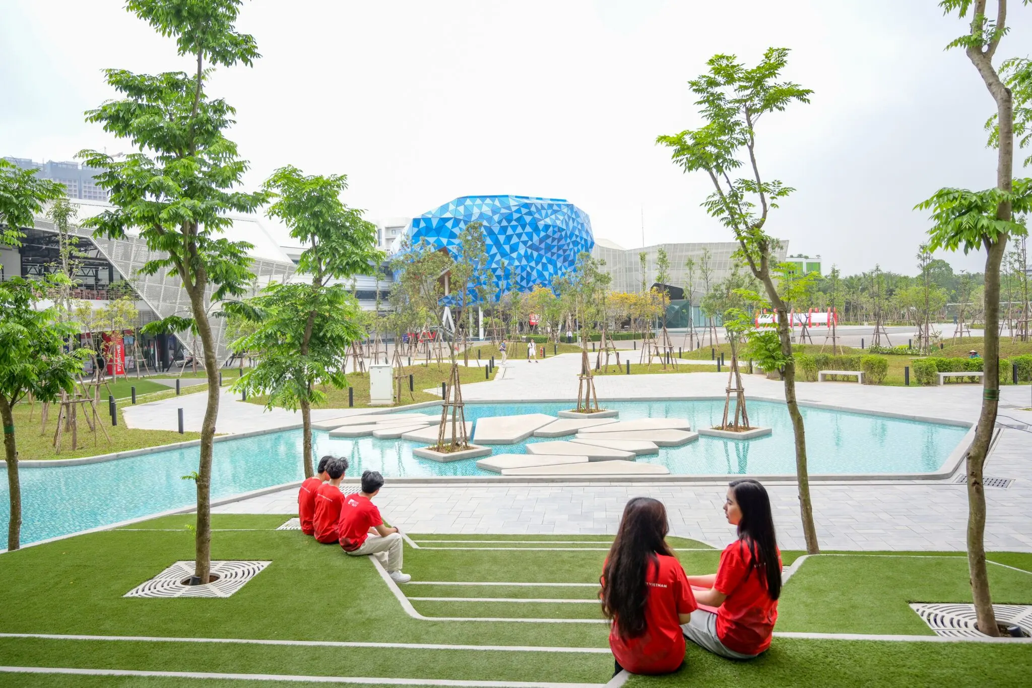 Four students in red shirts relax on a landscaped lawn overlooking a reflecting pool and the modern BUV campus.