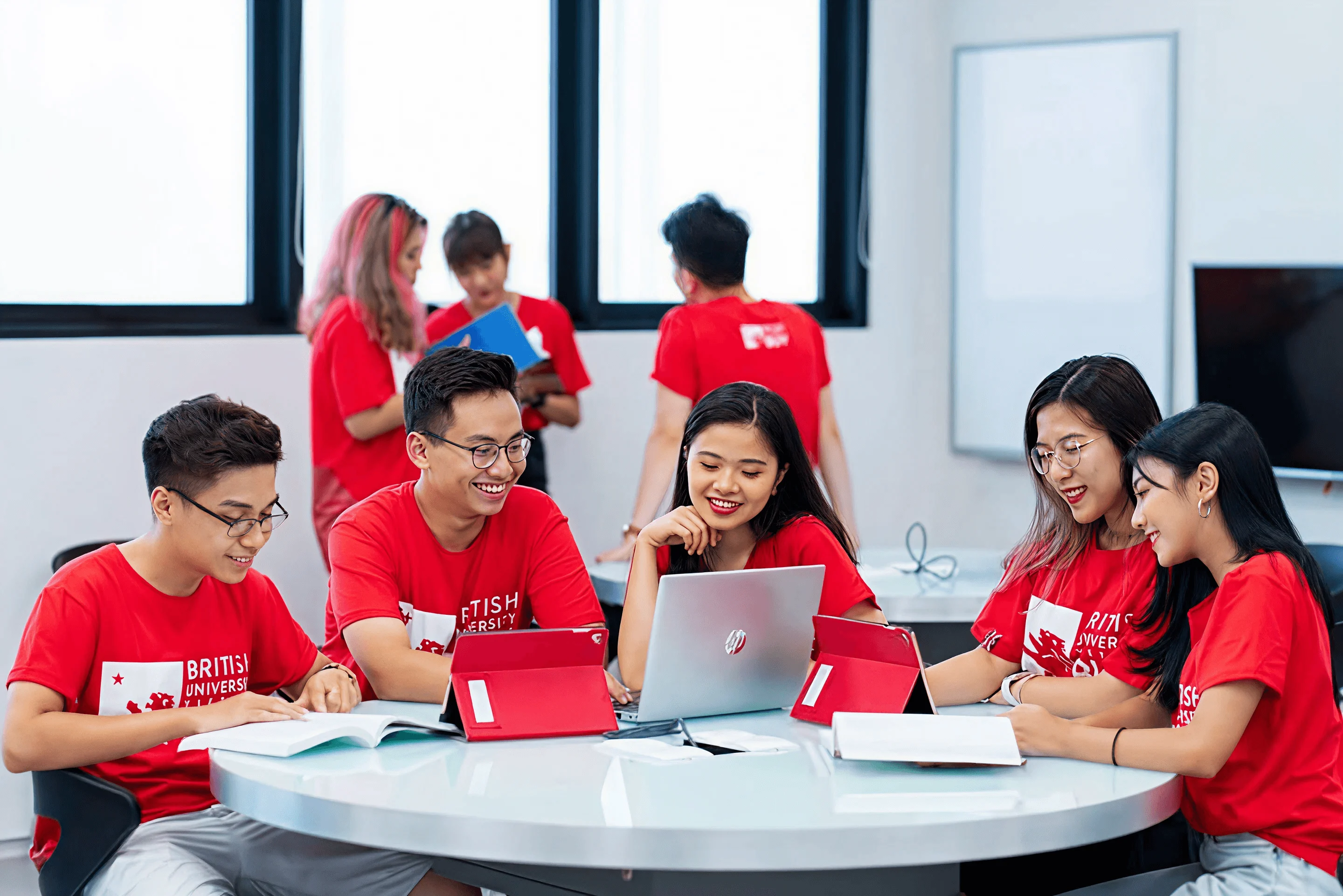 BUV students in red shirts collaborate around a table using laptops and tablets in a bright classroom.
