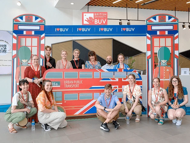 A group of people posing in front of a British-themed London bus display at British University Vietnam, with “I ♥ BUV” and BUV signs visible.