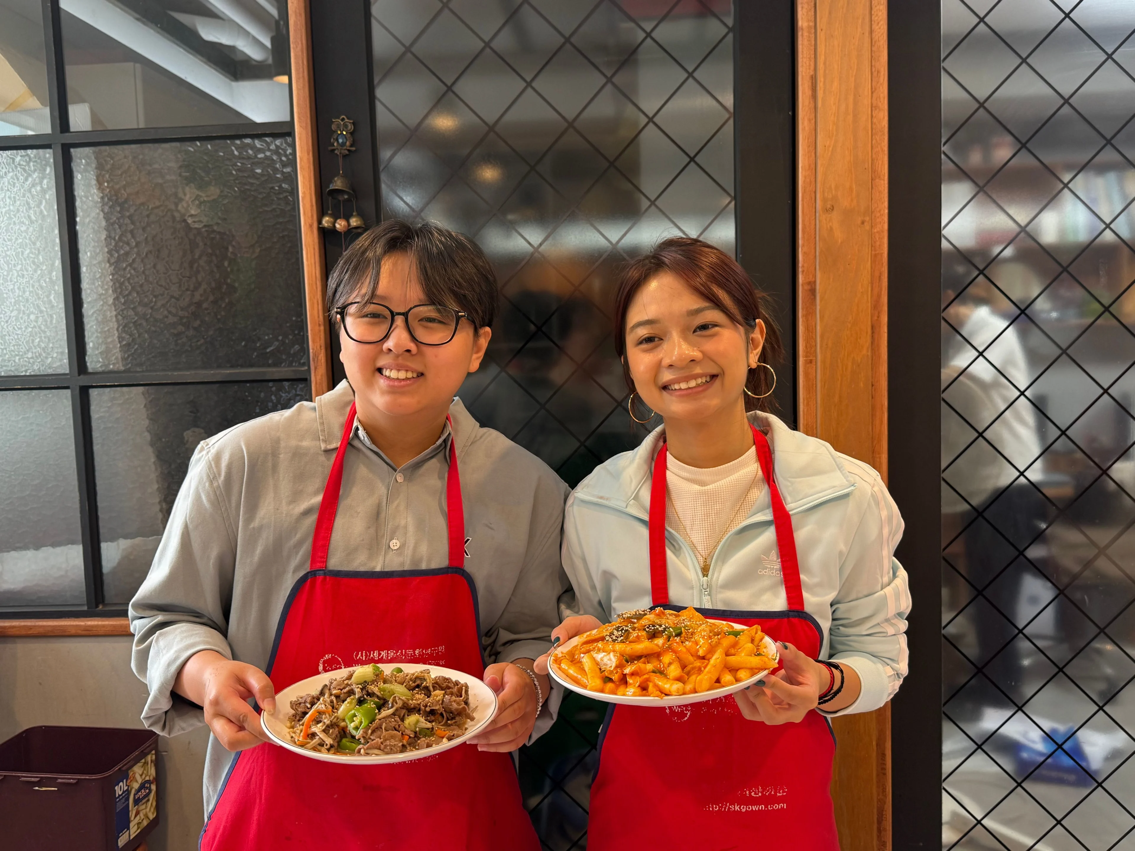 Two students wearing red aprons smile while holding plates of food during a cultural cooking experience.