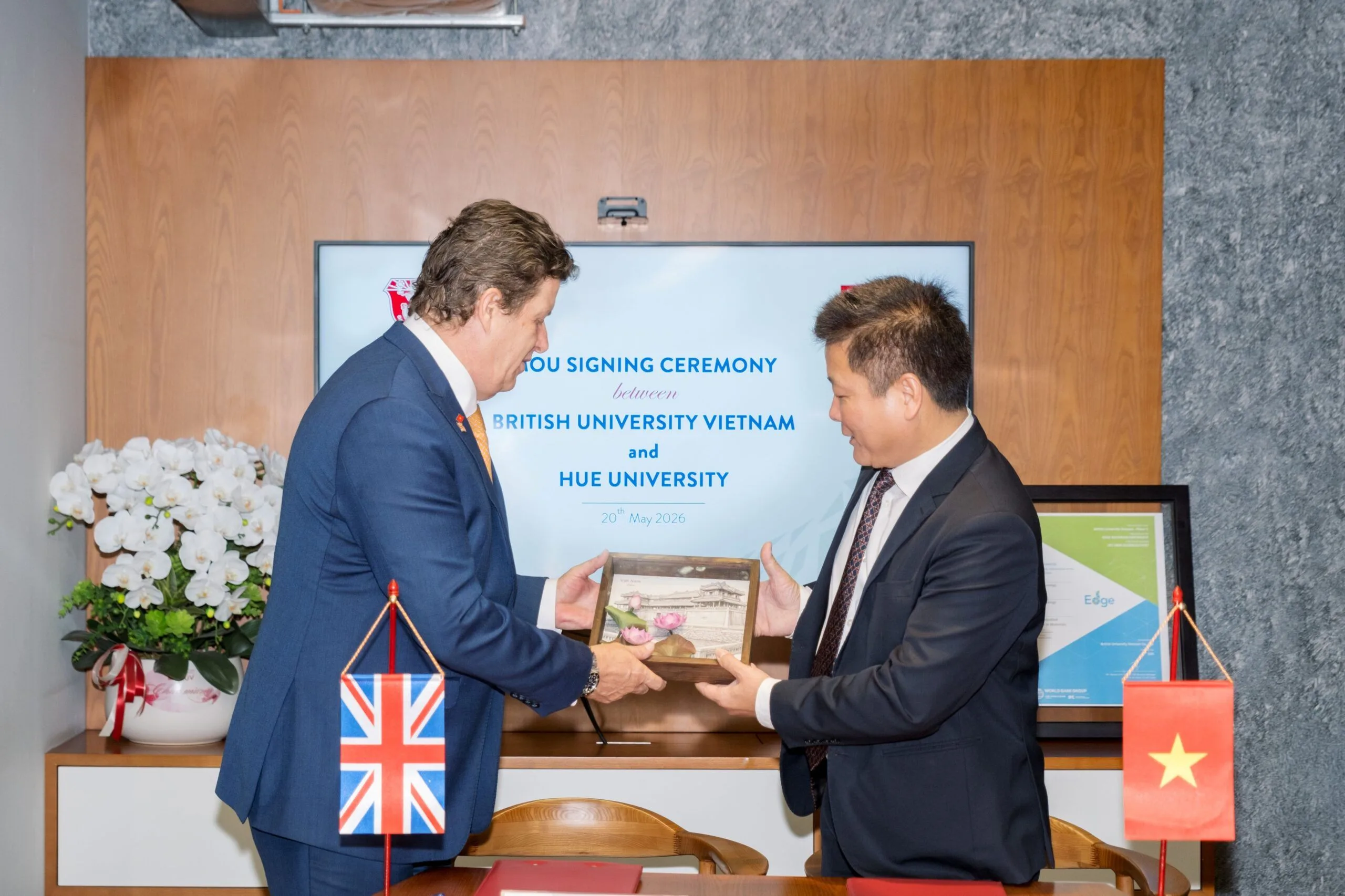 Two men in suits exchange a signed document during a university signing ceremony in front of a presentation backdrop, with national flags of the UK and Vietnam visible.