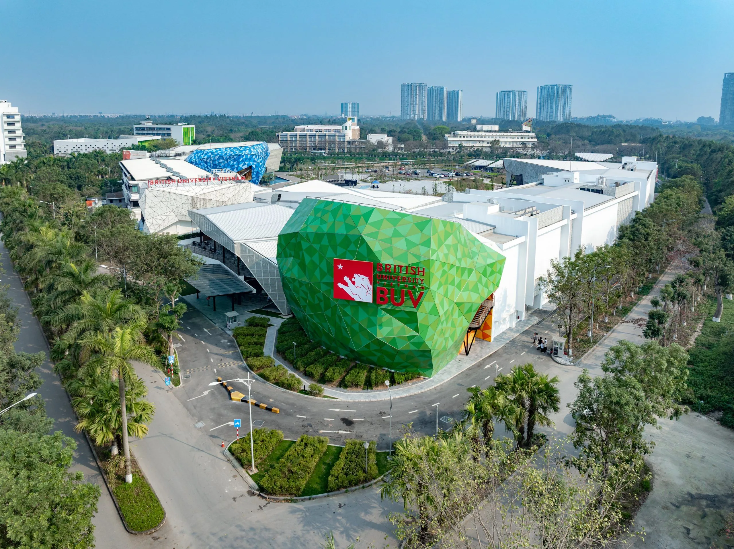 Aerial view of the British University Vietnam campus, featuring a distinctive green geometric façade with the red BUV logo, surrounded by tree-lined roads.