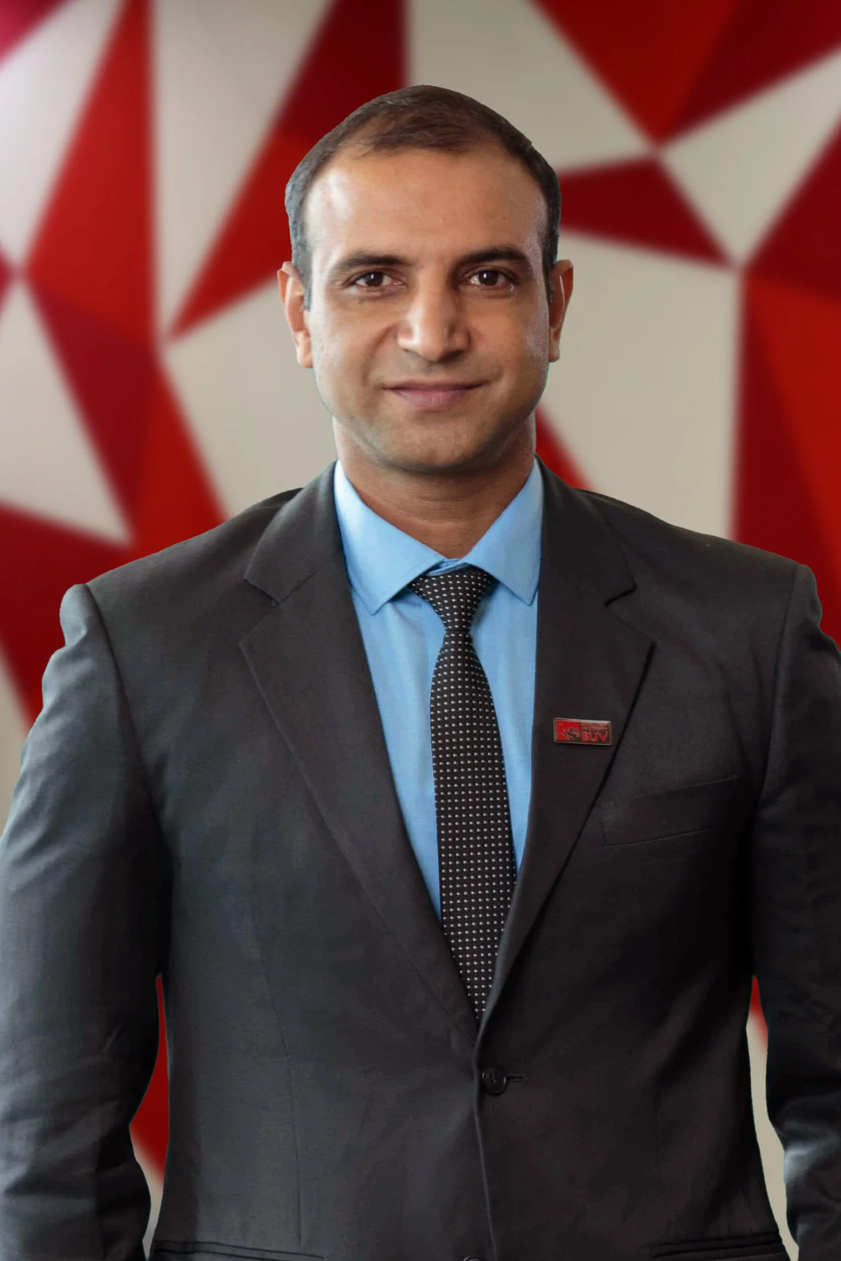 Alt text: Portrait of a man in a dark suit and blue shirt with a red tie, standing in front of a red geometric backdrop.