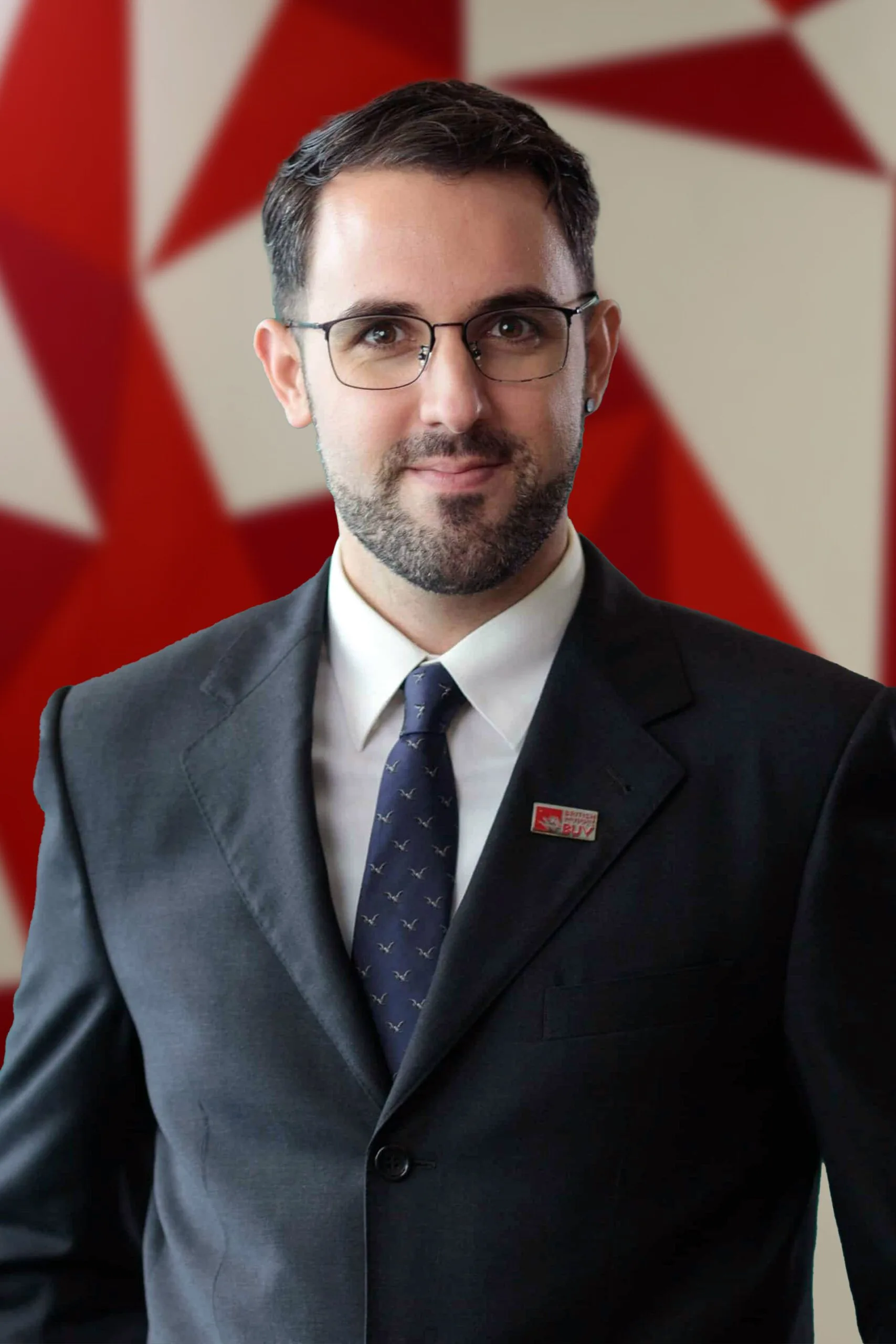 A man in a dark suit and tie wearing glasses, posing for a formal portrait against a red-and-white geometric background.