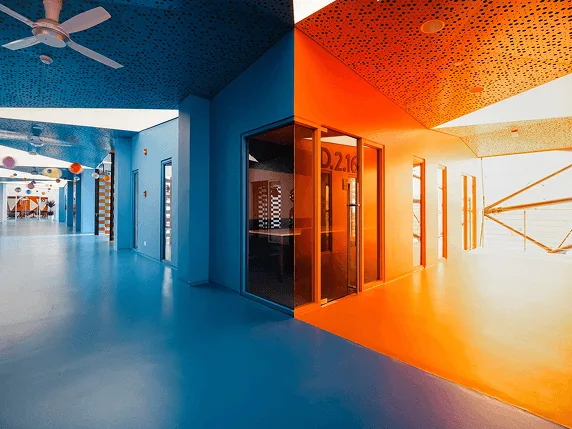 Modern university campus interior with blue and orange corridors, glass doors, ceiling fans, and bright geometric lighting.