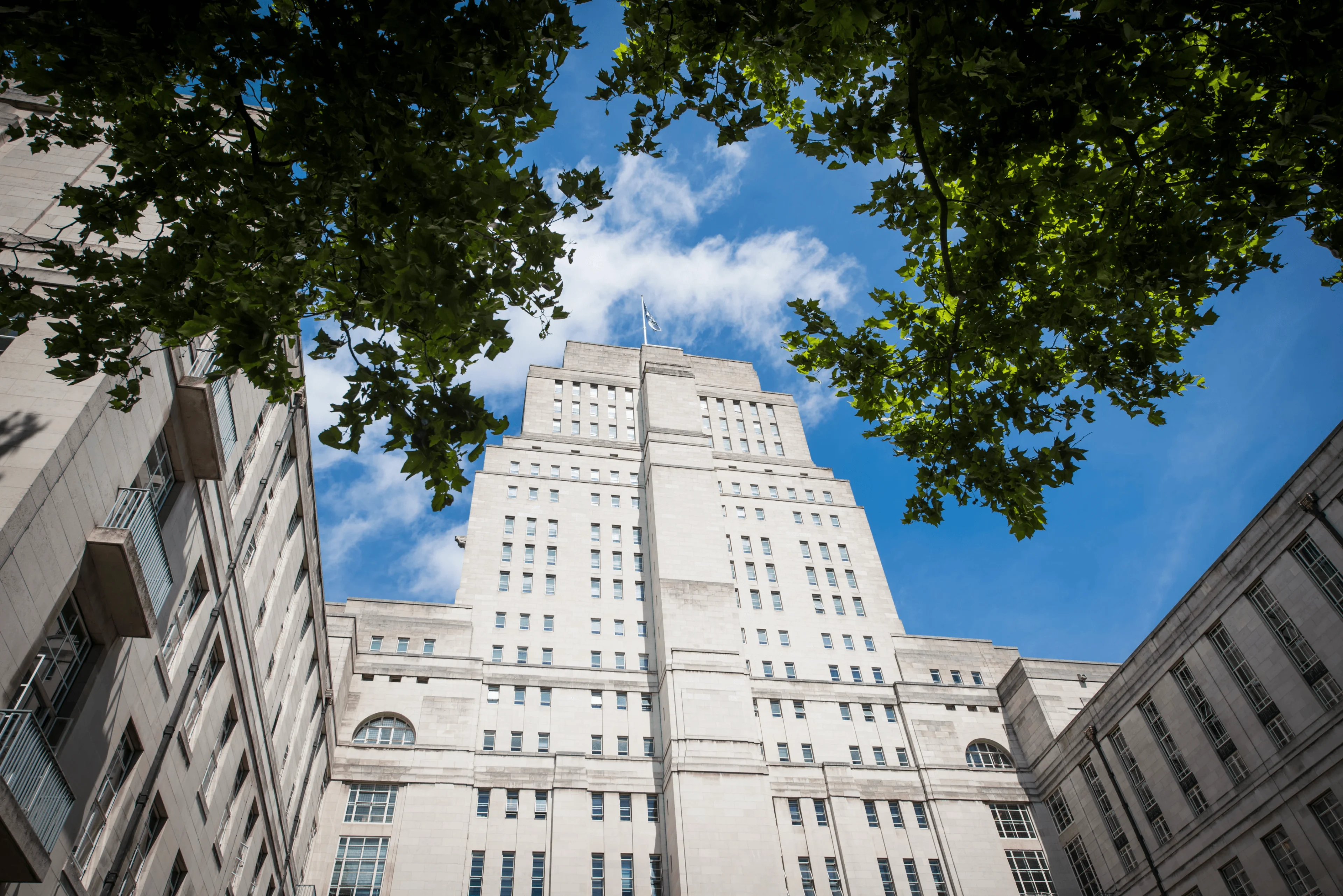Low-angle view of the University of London’s Senate House framed by leafy trees against a blue sky.