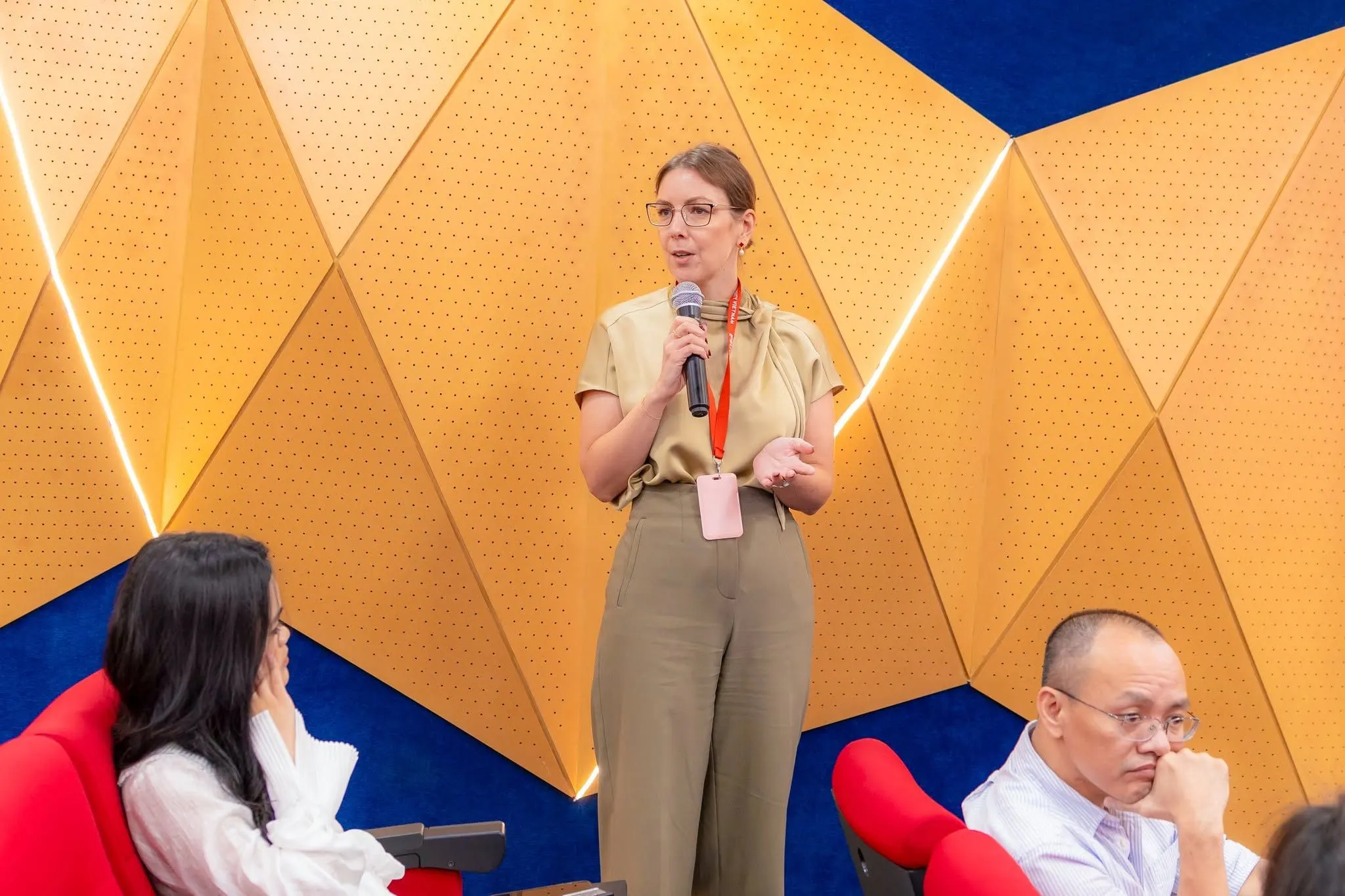 A woman speaks into a microphone during an event, with two attendees seated in front of a geometric wooden wall.