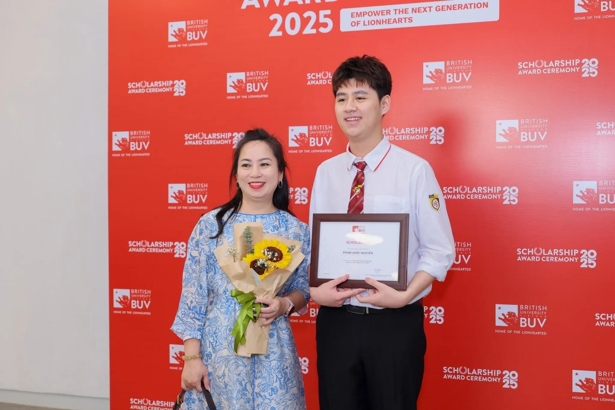 A BUV Scholarship Award 2025 recipient holds a framed certificate while posing with a woman carrying a bouquet in front of a red BUV backdrop.
