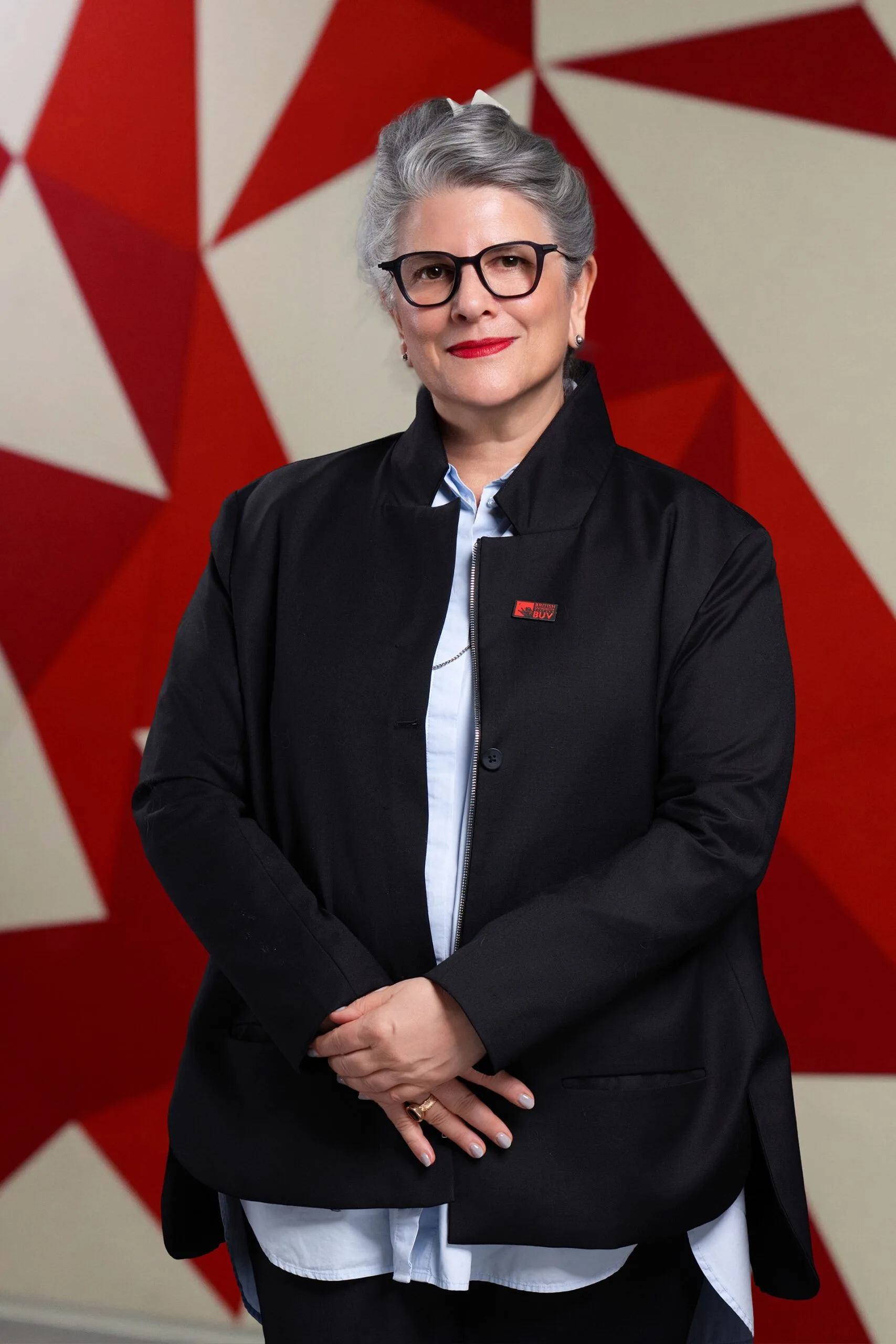 Sandra Schneiderman wearing glasses and a black BUV blazer against a red geometric background
