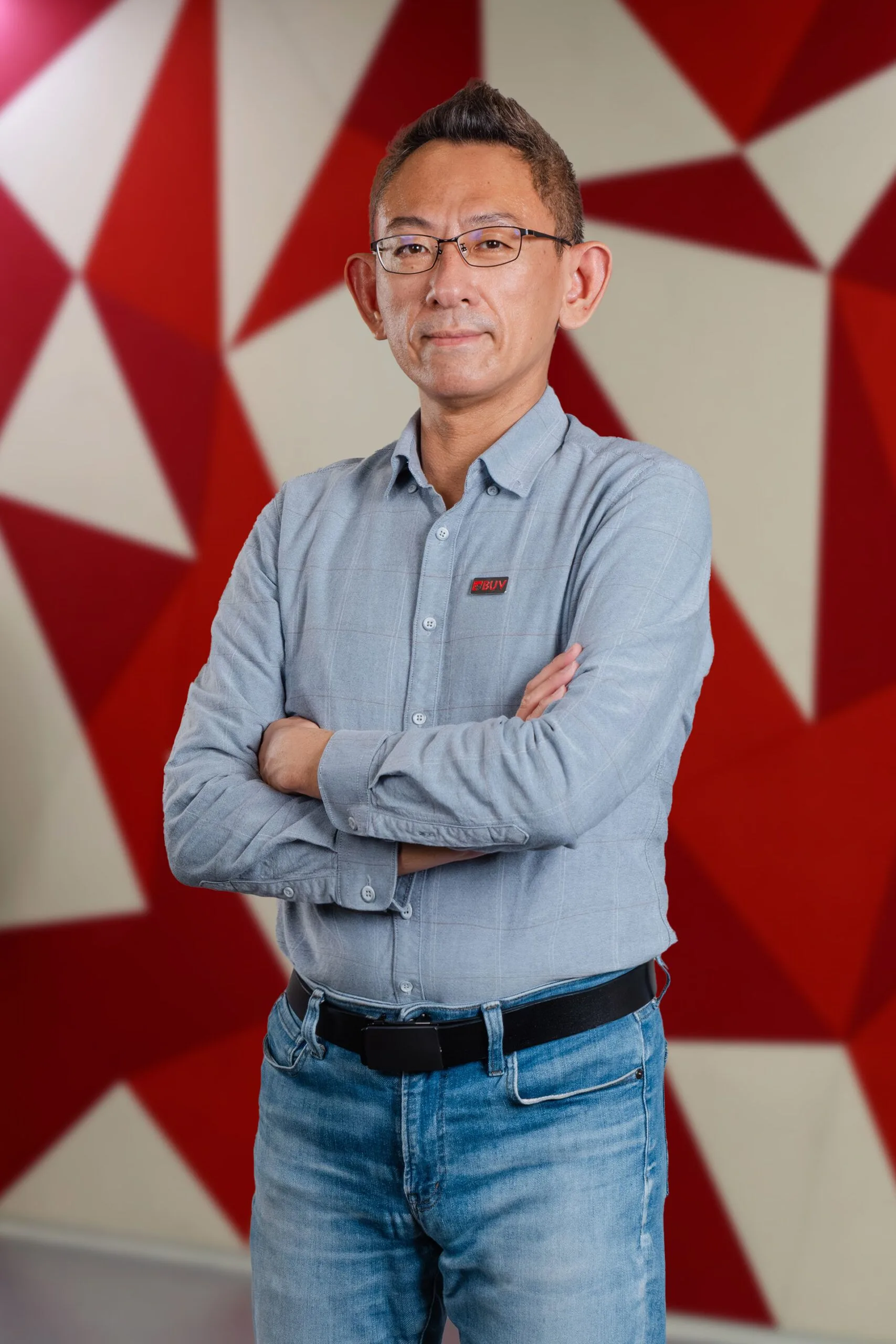 Portrait of a BUV staff member wearing glasses and a light blue BUV shirt, standing with arms crossed against a red-and-white geometric background.