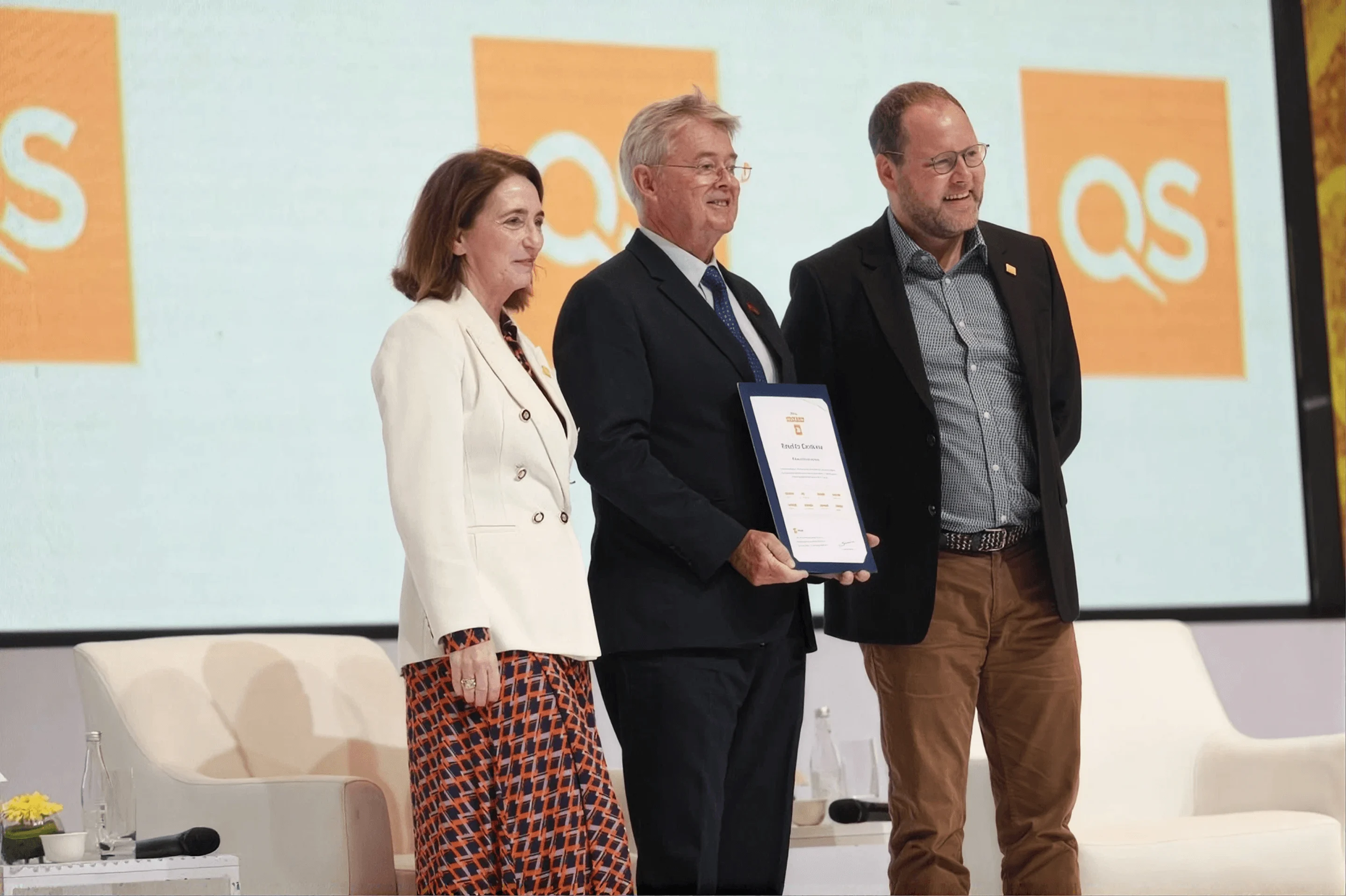 Three people on a stage presenting an official QS accreditation certificate in front of a backdrop featuring QS logos.