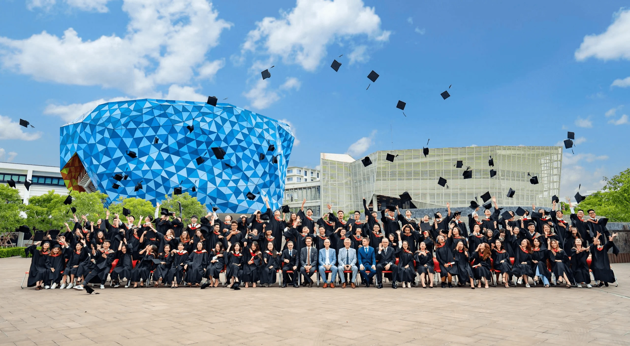 A large group of BUV graduates in caps and gowns celebrate outdoors, tossing their mortarboards in front of the university’s distinctive blue campus building.