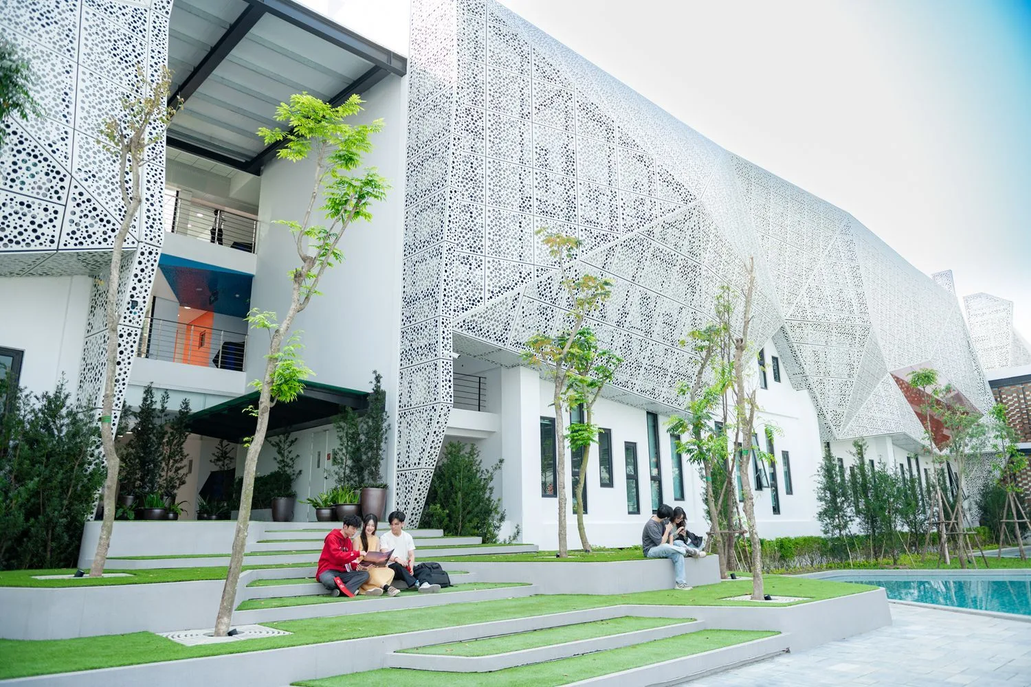 Sinh viên BUV học tập ngoài trời trong khuôn viên đại học hiện đại với kiến trúc mặt tiền đục lỗ và cây xanh.
