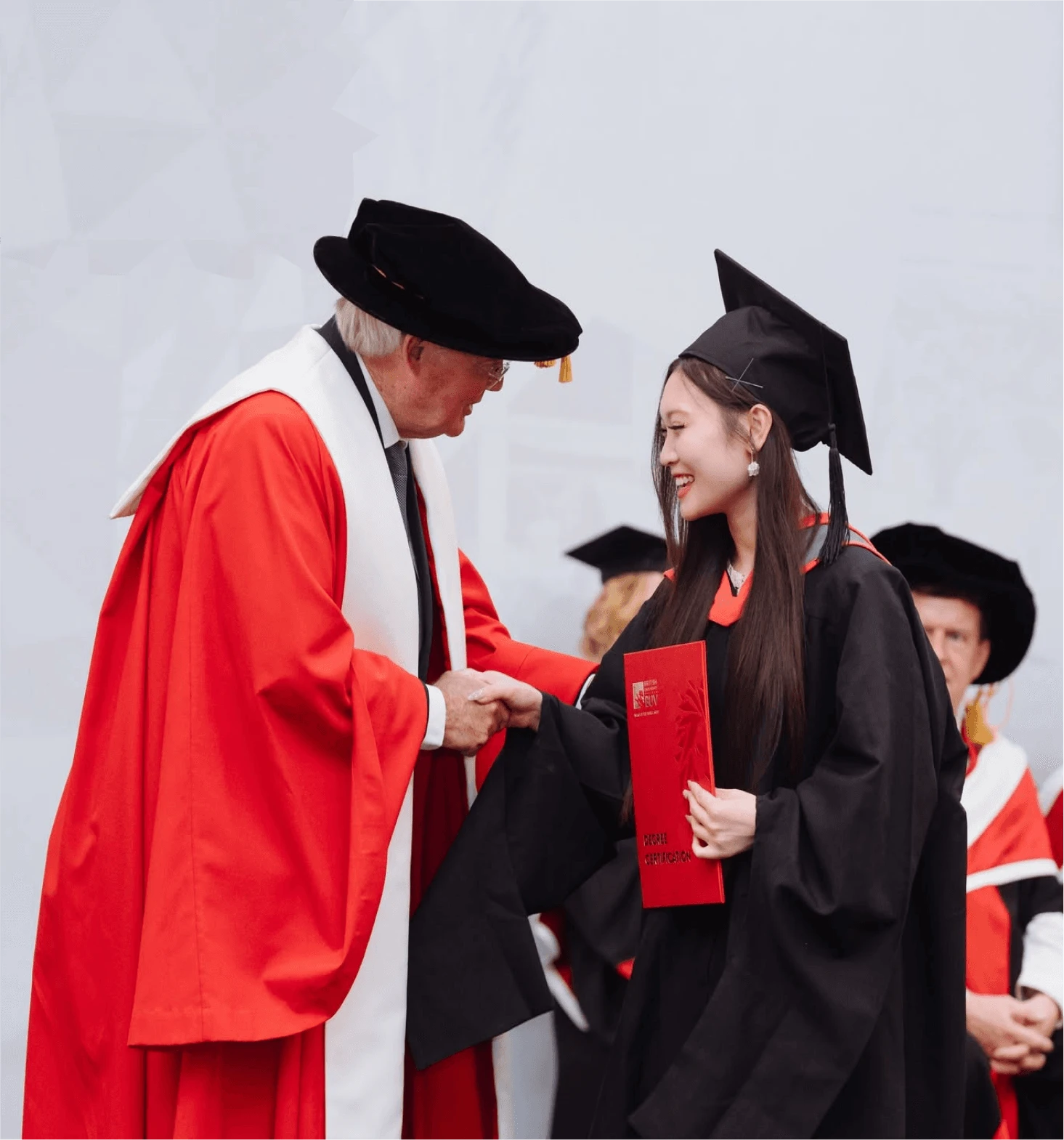 A smiling BUV graduate in cap and gown shakes hands with a university official while holding her red diploma folder at a graduation ceremony.