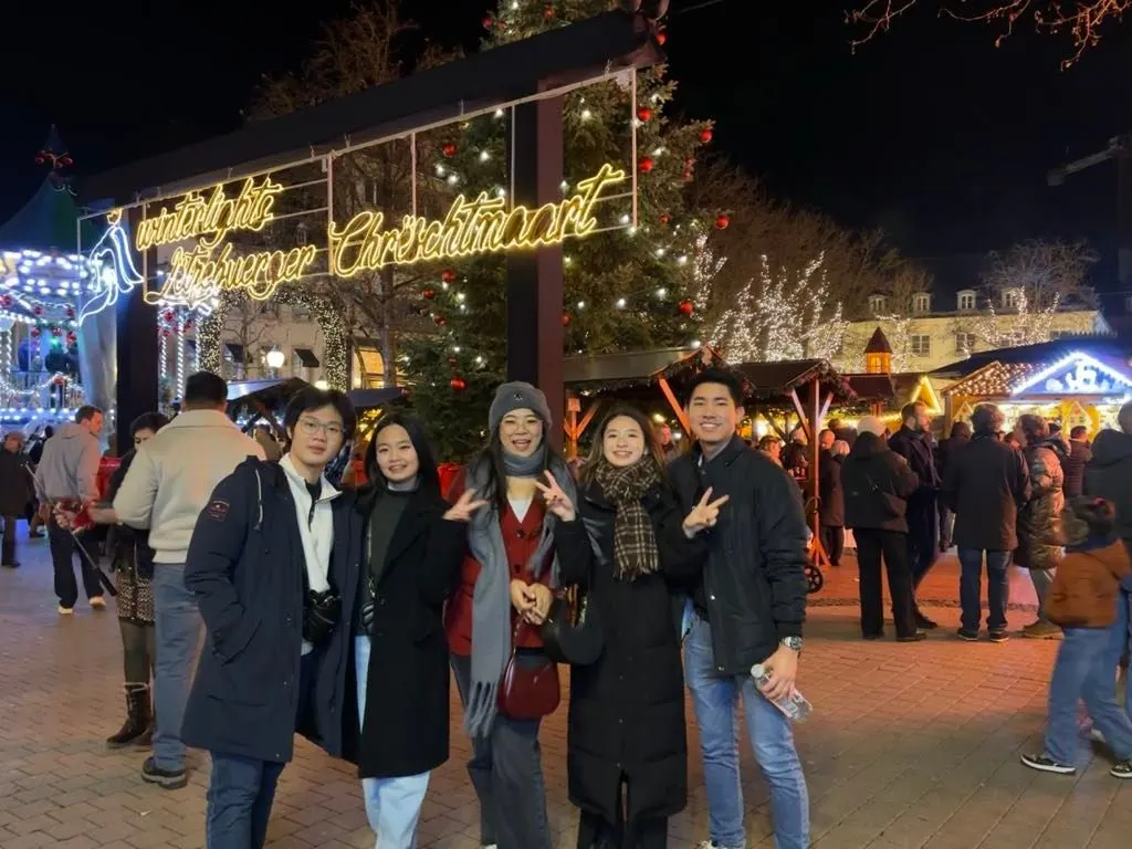 Five students pose together at a brightly lit Christmas market at night.