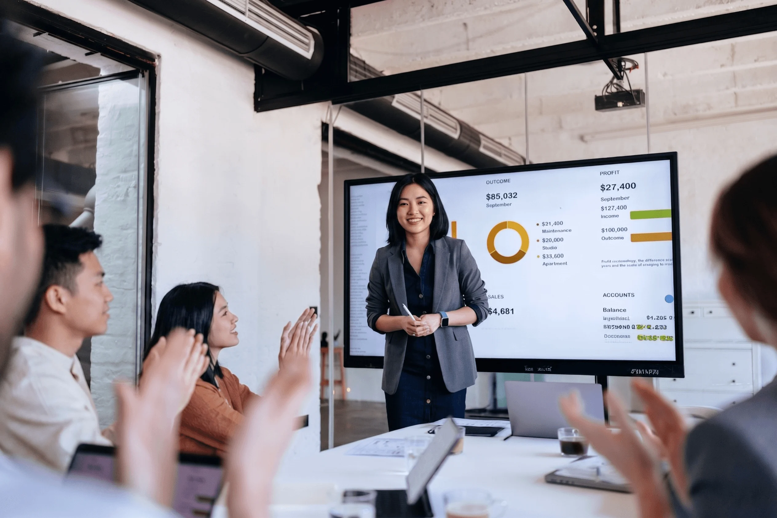 A businesswoman presents financial data on a screen as colleagues applaud in a modern meeting room.