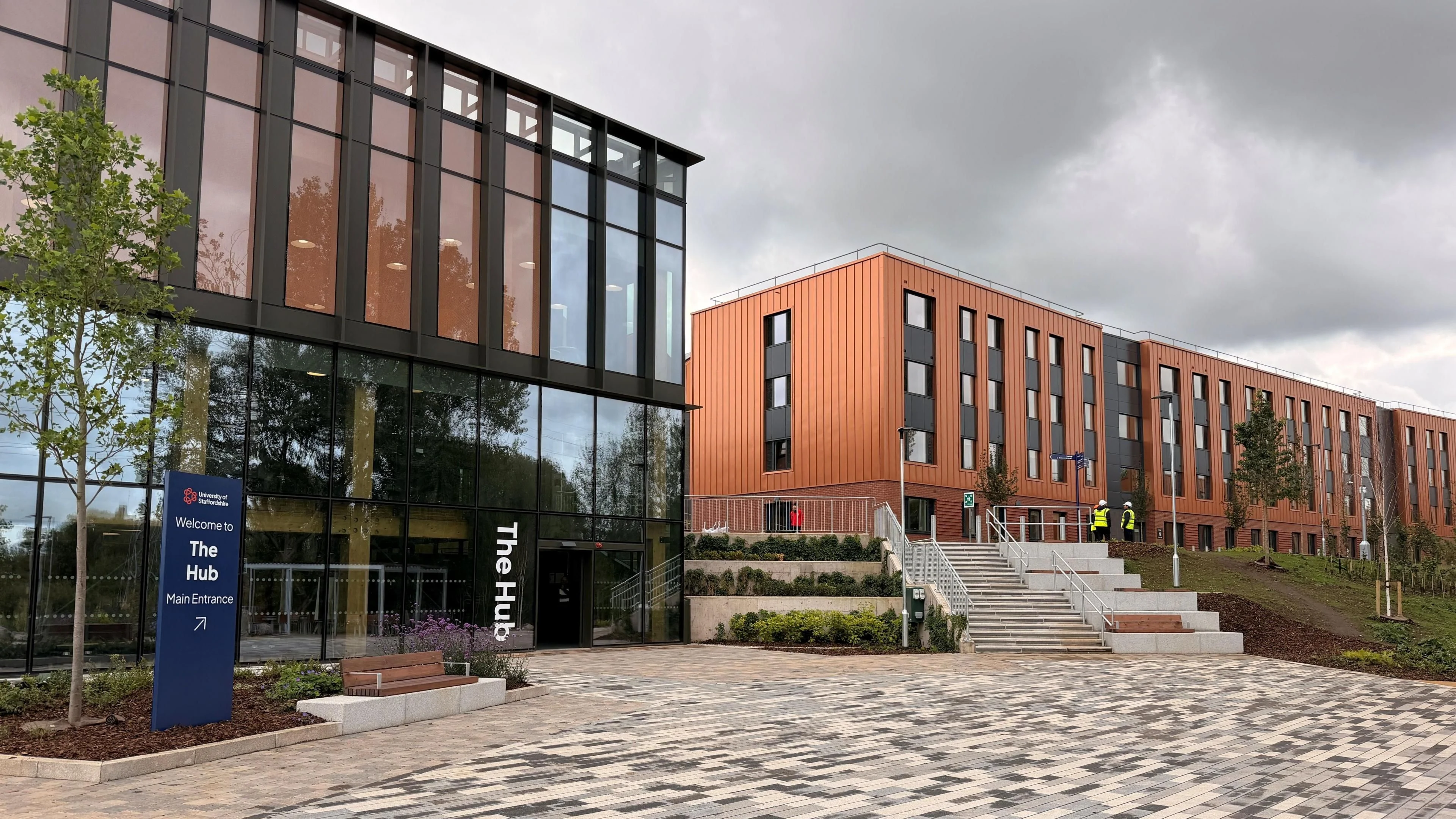 An exterior shot of a modern, multi-story brick and glass building identified by a sign as "The Hub" at Staffordshire University. Large windows dominate the building's facade, reflecting the cloudy sky. A set of concrete stairs and a paved walkway lead up to the entrance. Landscaping elements like small trees and a wooden bench are also visible. In the background, there are other buildings with brick exteriors and a few individuals can be seen. The overall mood is businesslike and contemporary.