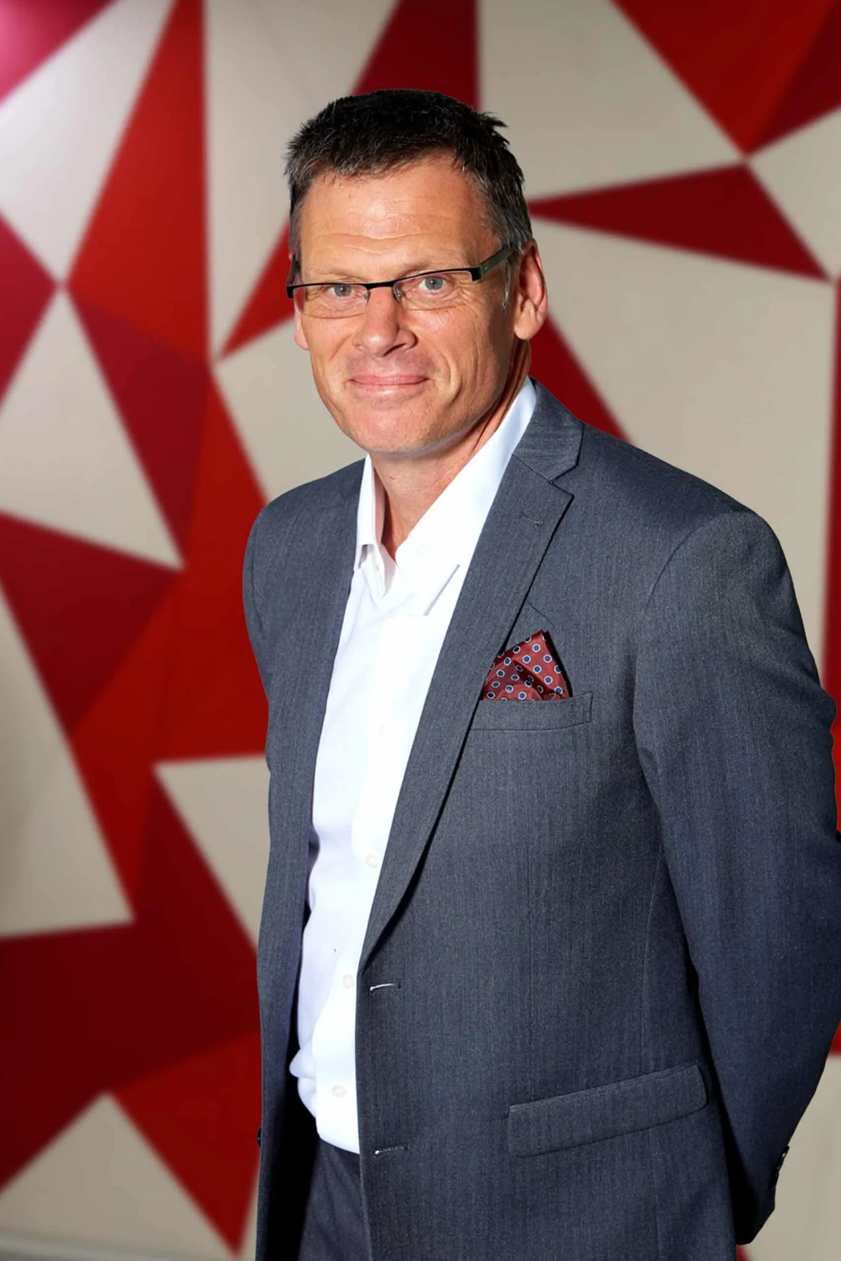 Portrait of Paul Gough in a blue suit and glasses, standing in front of a red-and-white geometric backdrop.