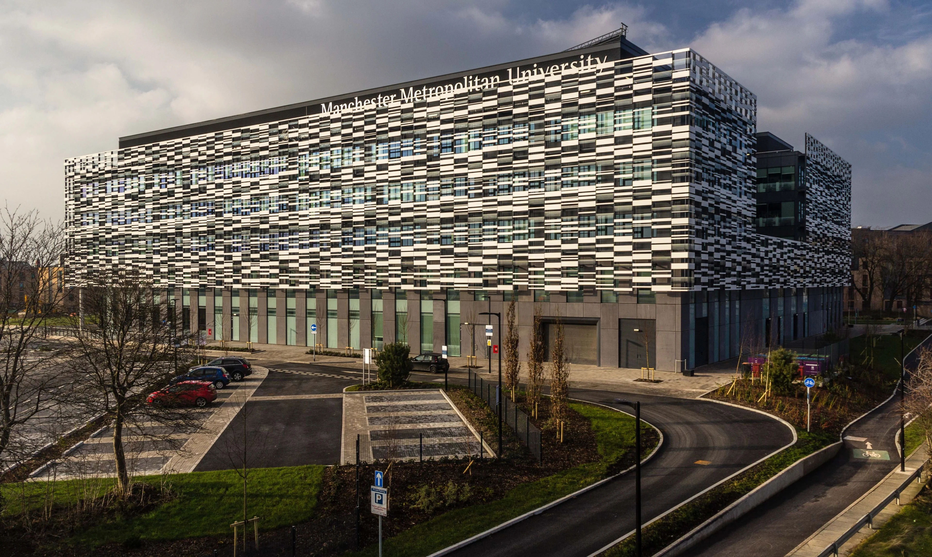 Modern Manchester Metropolitan University building with a patterned façade, viewed across campus roads and landscaped grounds.