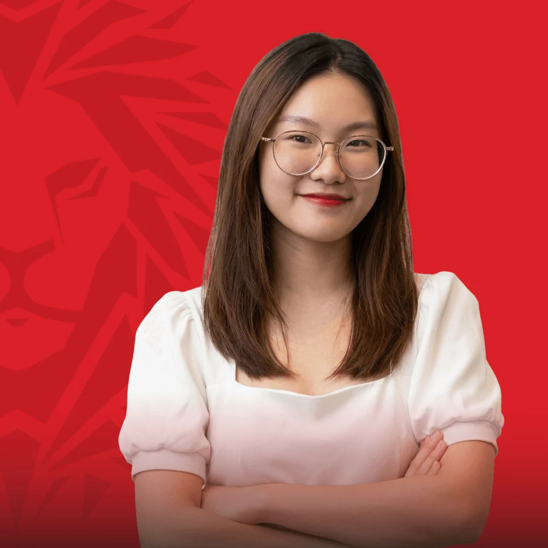 Portrait of a smiling female BUV alumna wearing glasses and a white blouse, standing with her arms crossed against a red background.