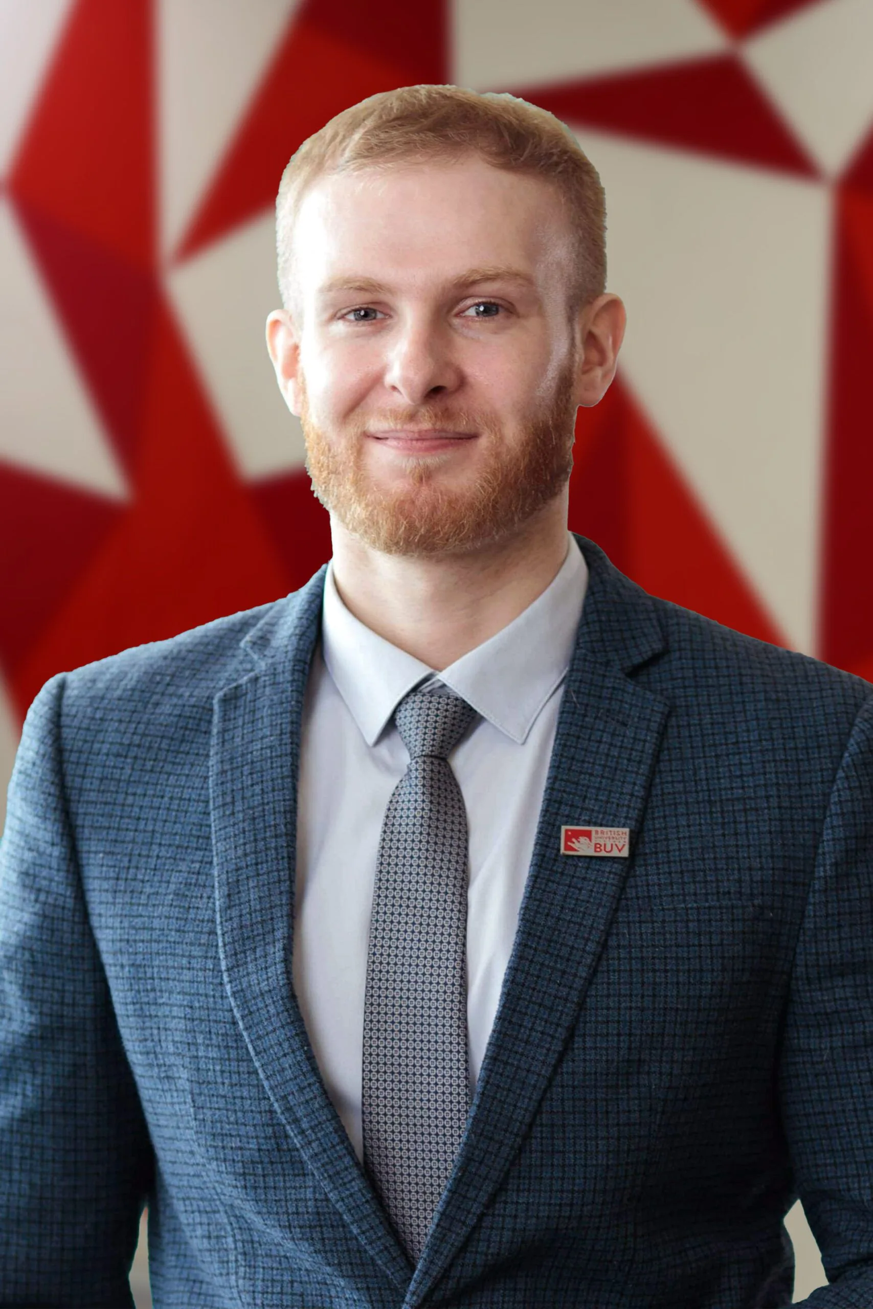 Portrait of a bearded man in a blue checked suit, white shirt and patterned tie, wearing a British University Vietnam (BUV) badge against a red-and-white geometric background.