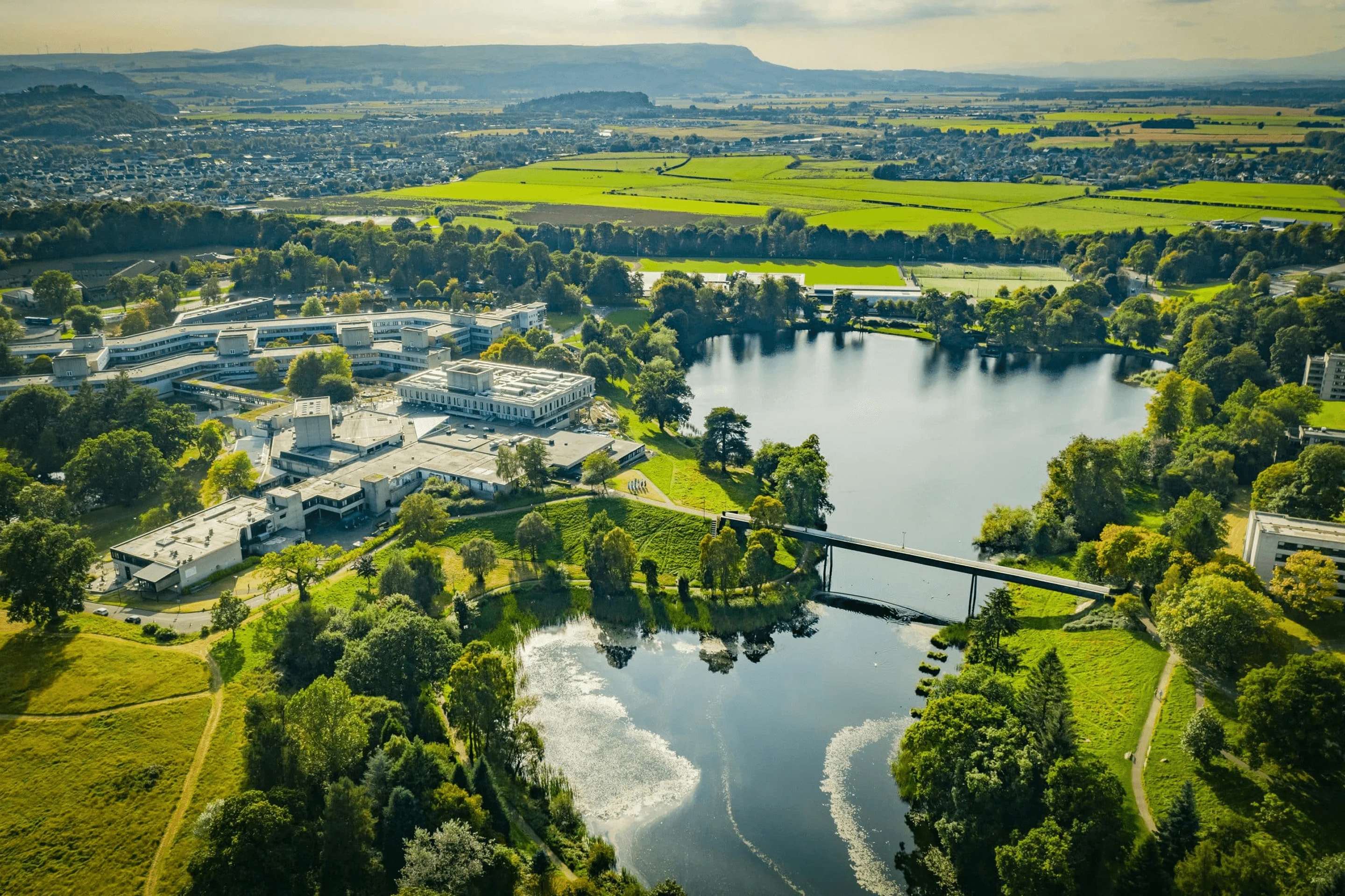 Aerial view of the University of Stirling campus, surrounded by a lake, lush greenery, and countryside.