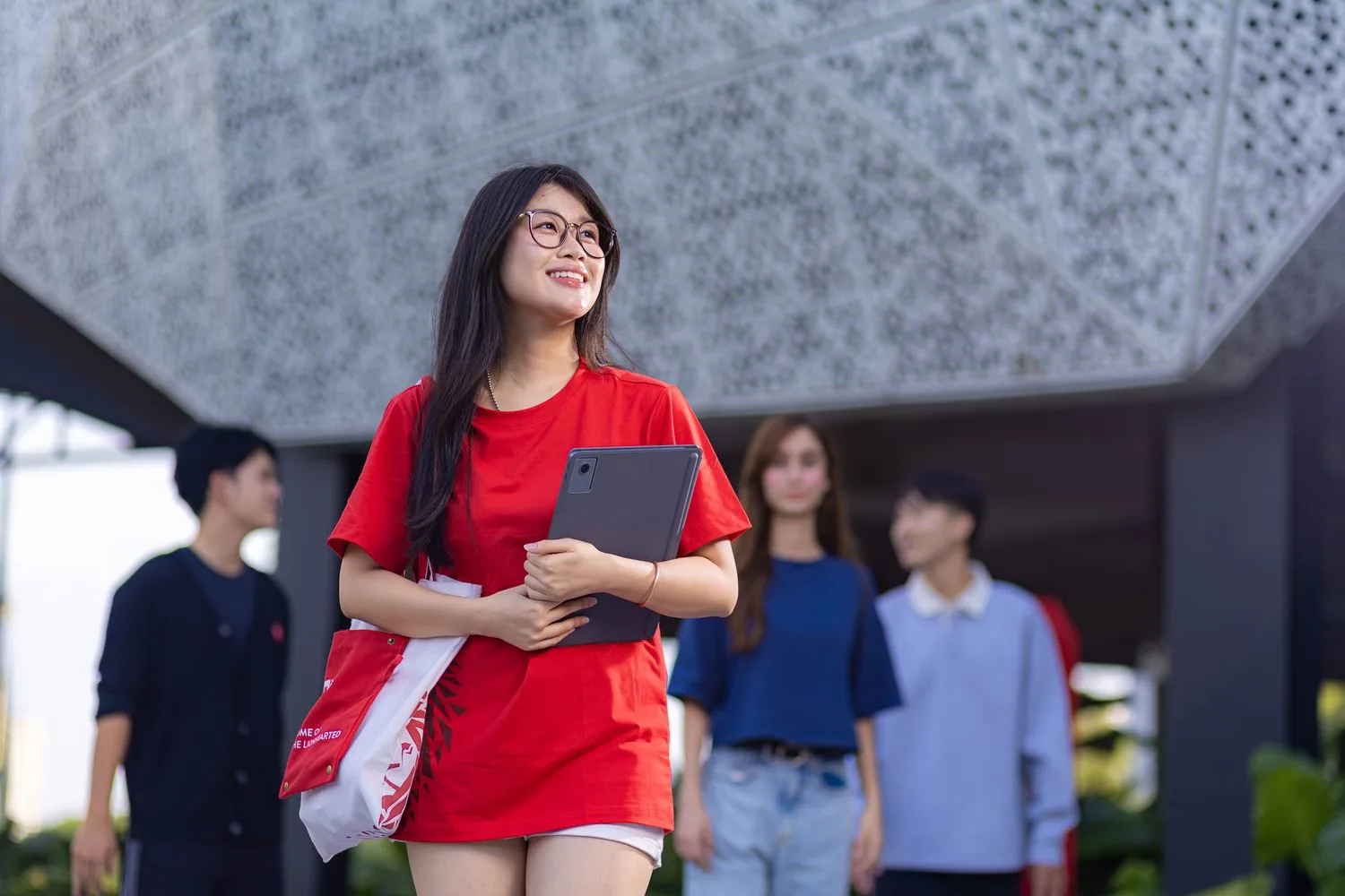BUV student in a red shirt holding a tablet on campus with fellow students in the background.