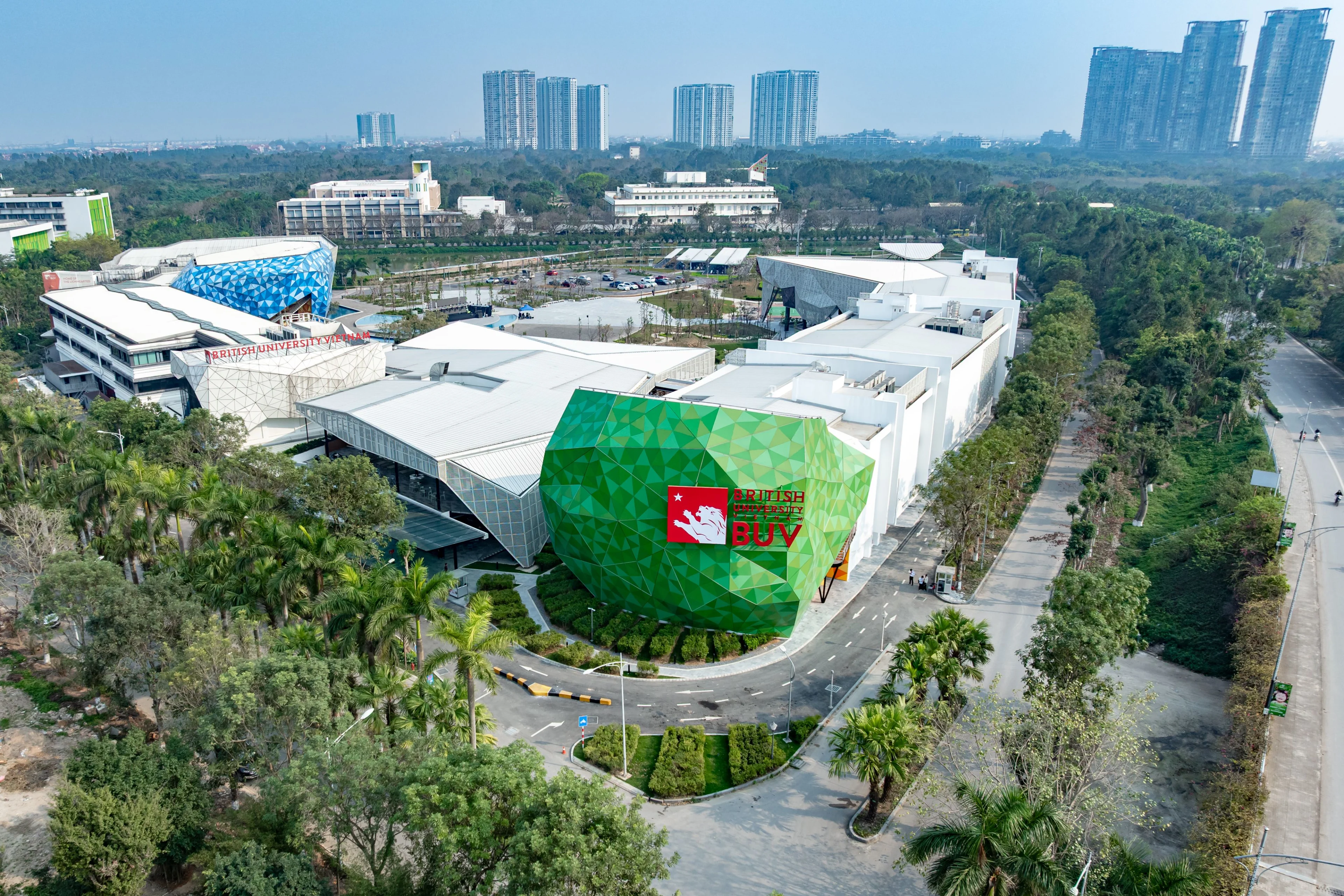 Aerial view of British University Vietnam (BUV) campus, featuring colorful geometric buildings, landscaped grounds, palm trees, surrounding roads, and a city skyline in the background.