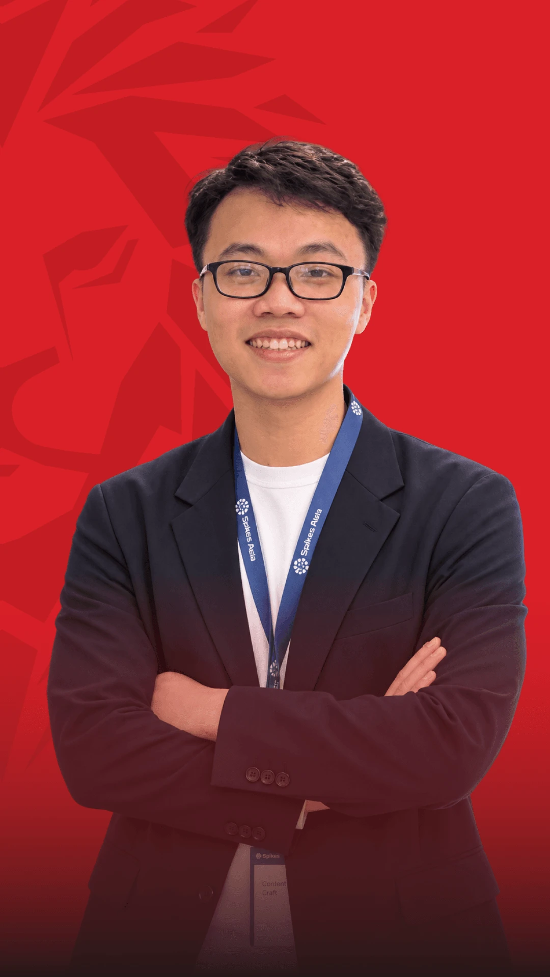Ảnh chân dung một sinh viên/cựu sinh viên nam đang bắt tay trước ngực, mặc áo vest sẫm màu, đeo thẻ thẻ tên, đứng trước phông nền đỏ.