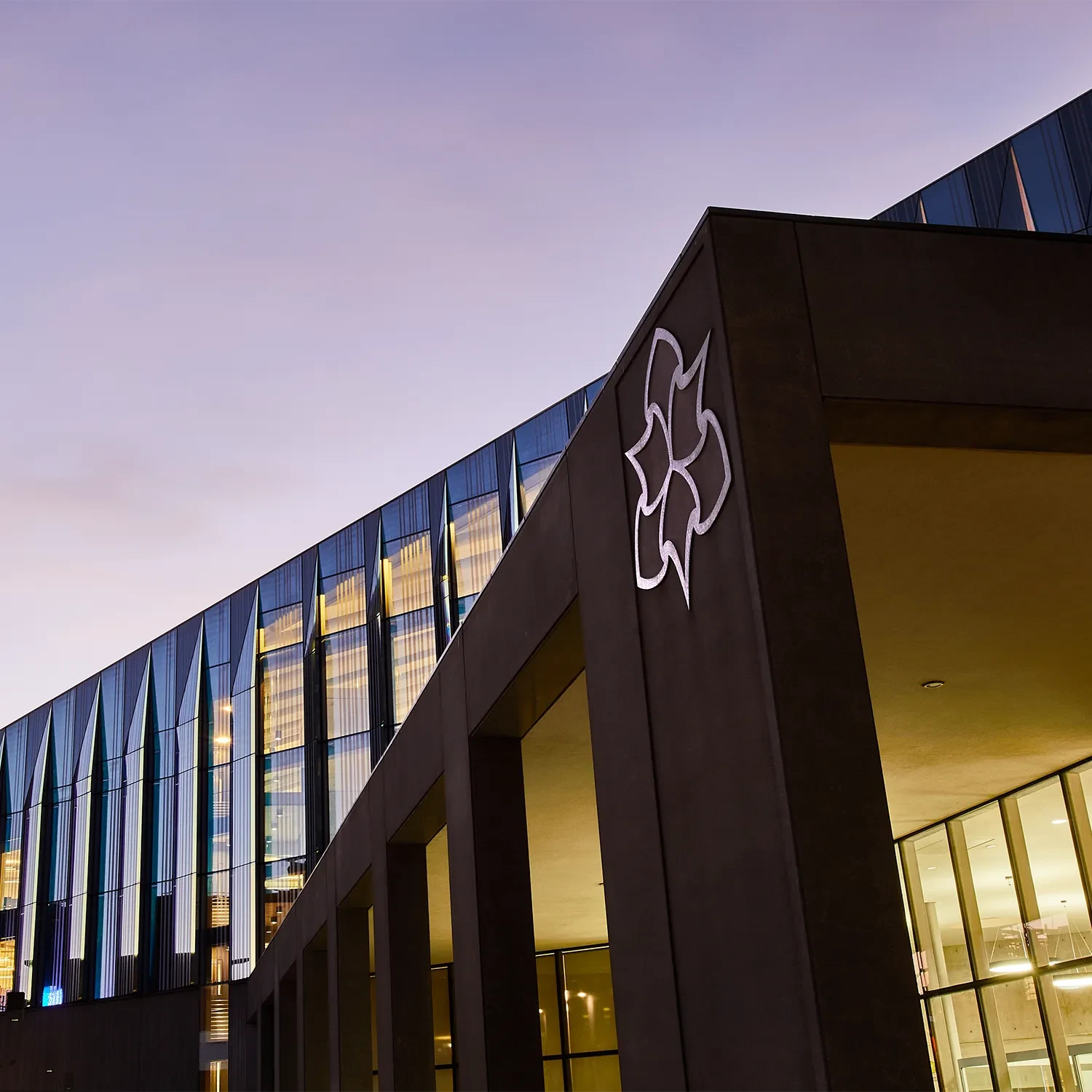 Low-angle view of a modern Manchester Metropolitan University building at dusk, highlighting its geometric glass facade and the illuminated university crest on an exterior pillar.