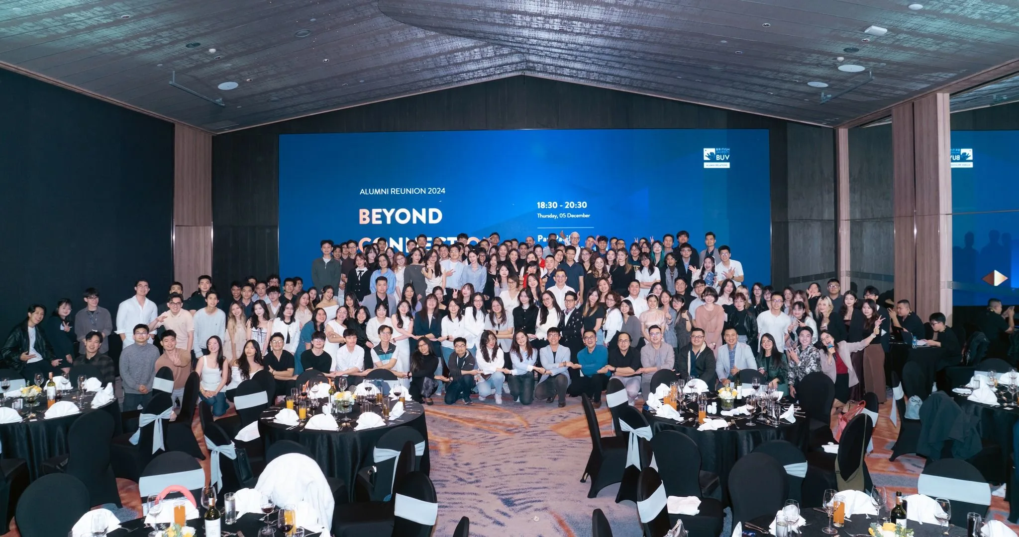A large group of attendees posing together for a photo in front of a stage backdrop reading "Alumni Reunion 2024" in a banquet hall.