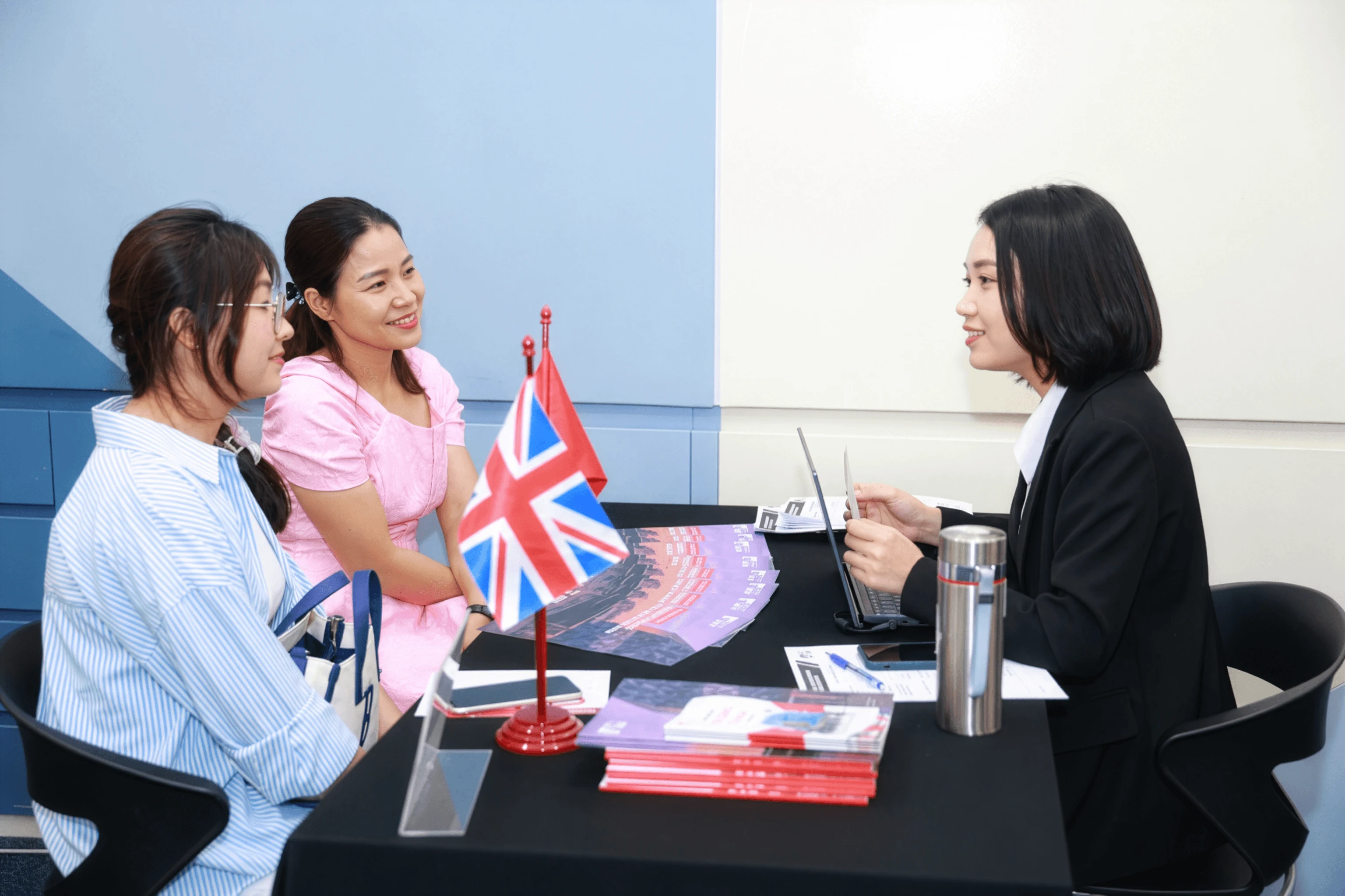 A university adviser speaks with two prospective students across a consultation table displaying a UK flag, brochures, a laptop, and stationery.
