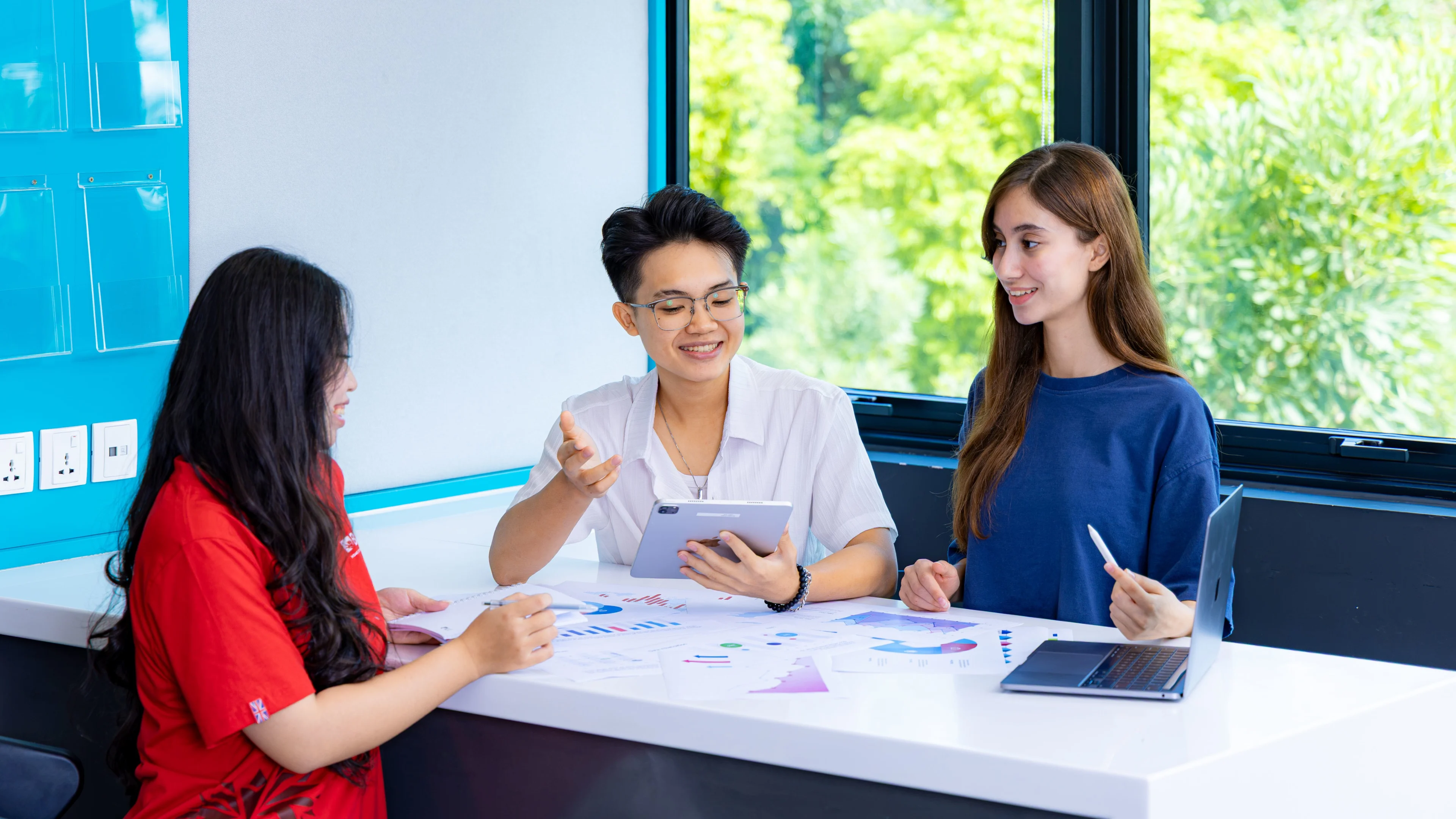 Three BUV students collaborate on coursework around a table with charts, a tablet and laptop in a bright study space.