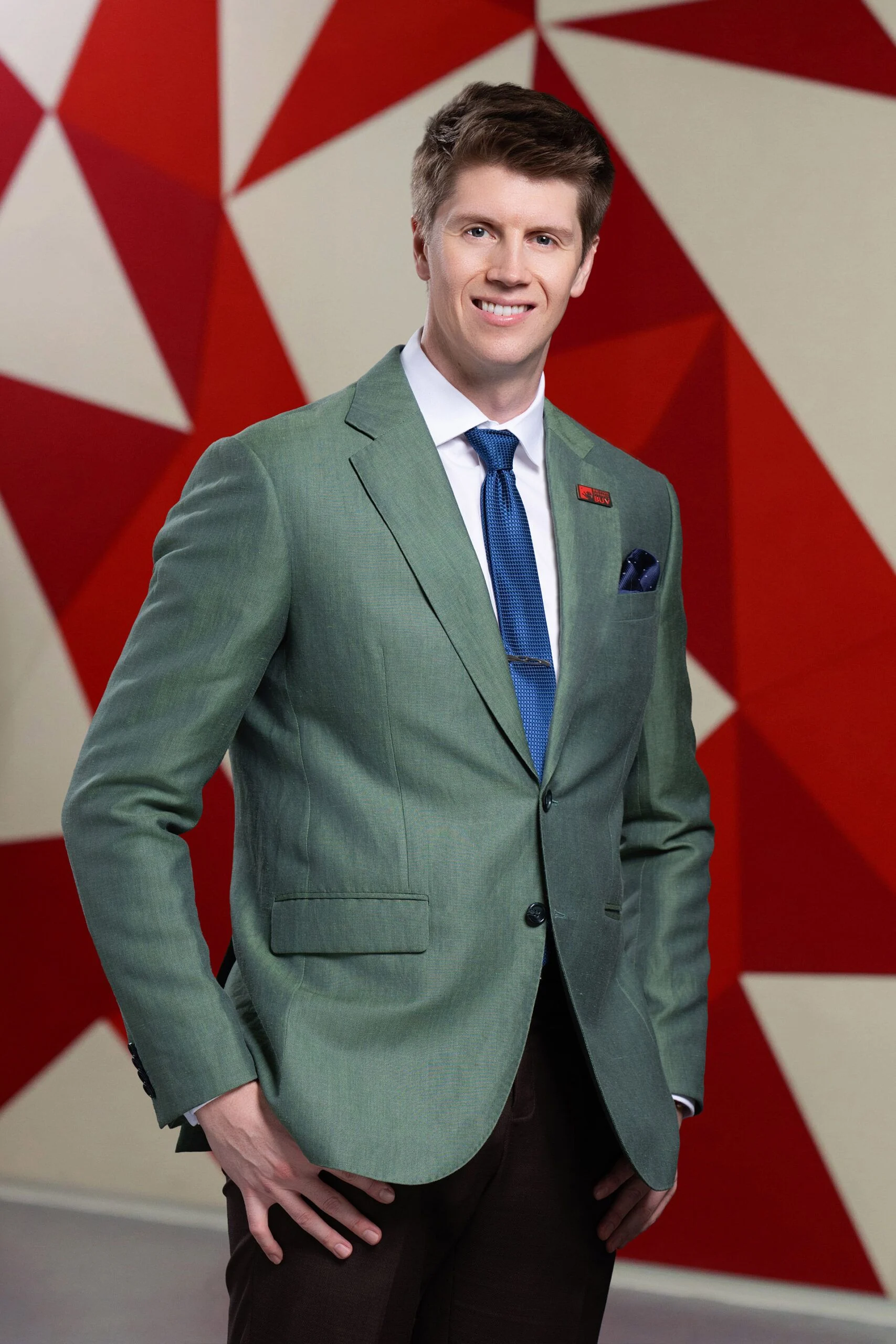 Mike Perkins smiles at the camera wearing a green suit, white shirt, and blue tie, standing in front of a red-and-white geometric background.