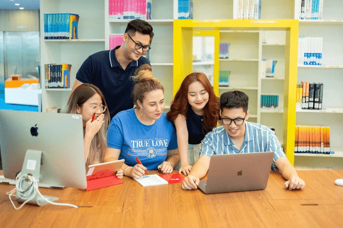 Five university students collaborate around a table with laptops and notebooks in a bright, colorful library.