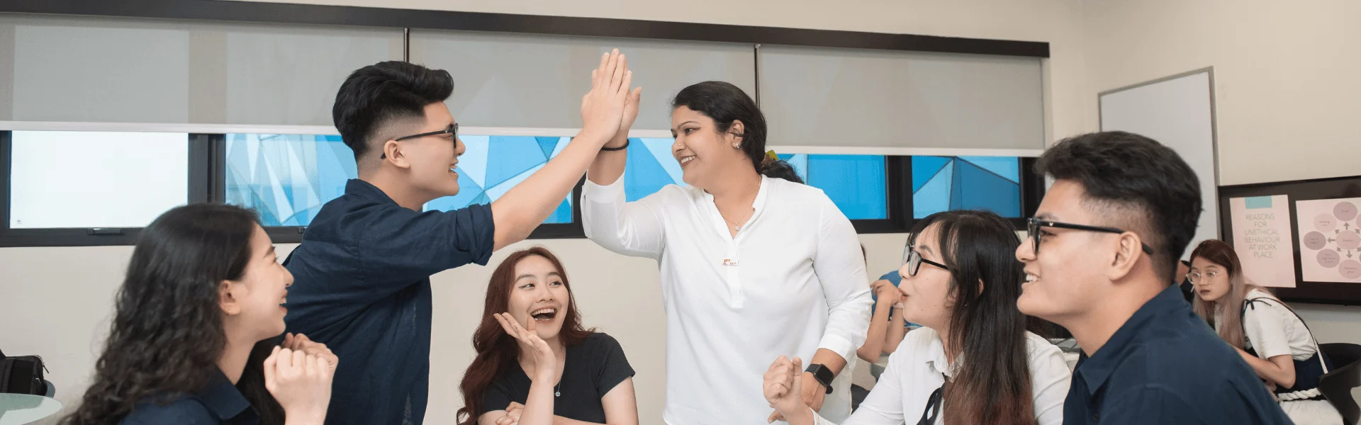 Alt text: A lecturer in a classroom greets a group of students, smiling and shaking hands while the students laugh and wave.