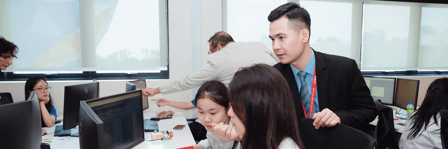 Students work at computers in a classroom while an instructor assists them.