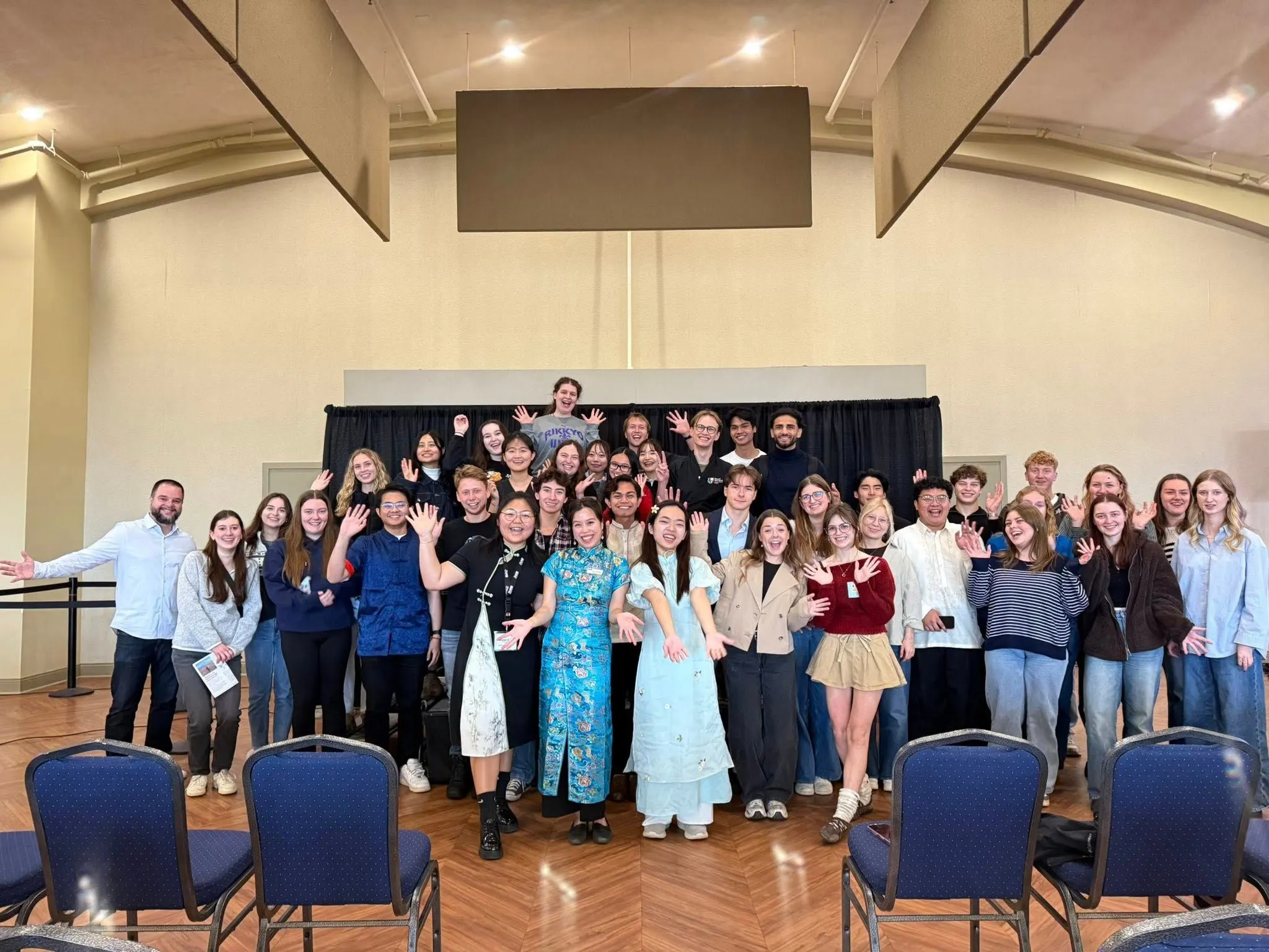 A diverse group of smiling students posing and waving together on a stage indoors, with several members at the front wearing traditional attire.